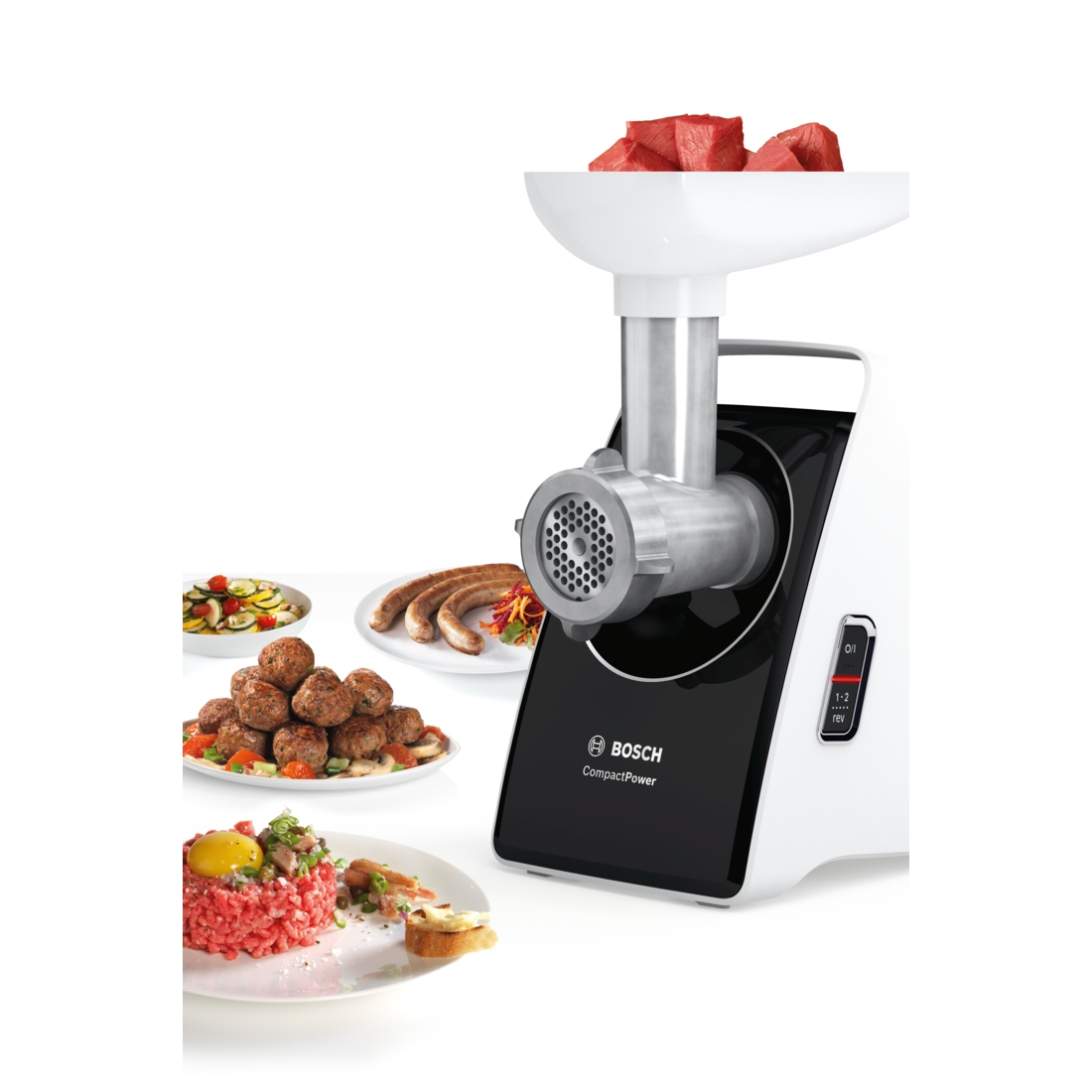 Un tritacarne Bosch bianco e nero. Carne rossa nel vassoio. Piatti di cibo: polpette, salsicce e tartare sono visibili.