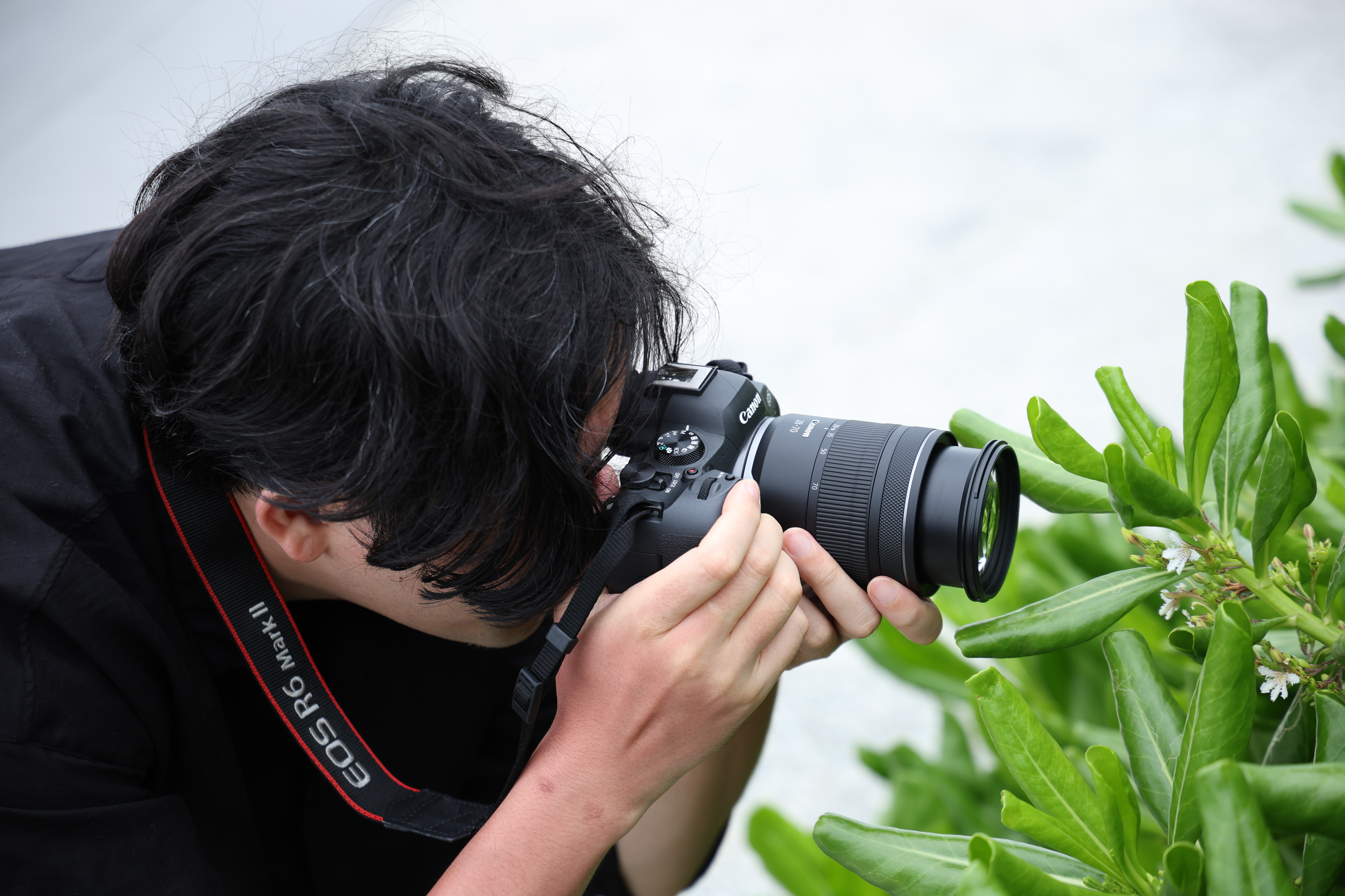 Siyah saçlı bir kişi Canon kamera ile fotoğraf çekiyor. Ön planda yeşil yapraklar var.