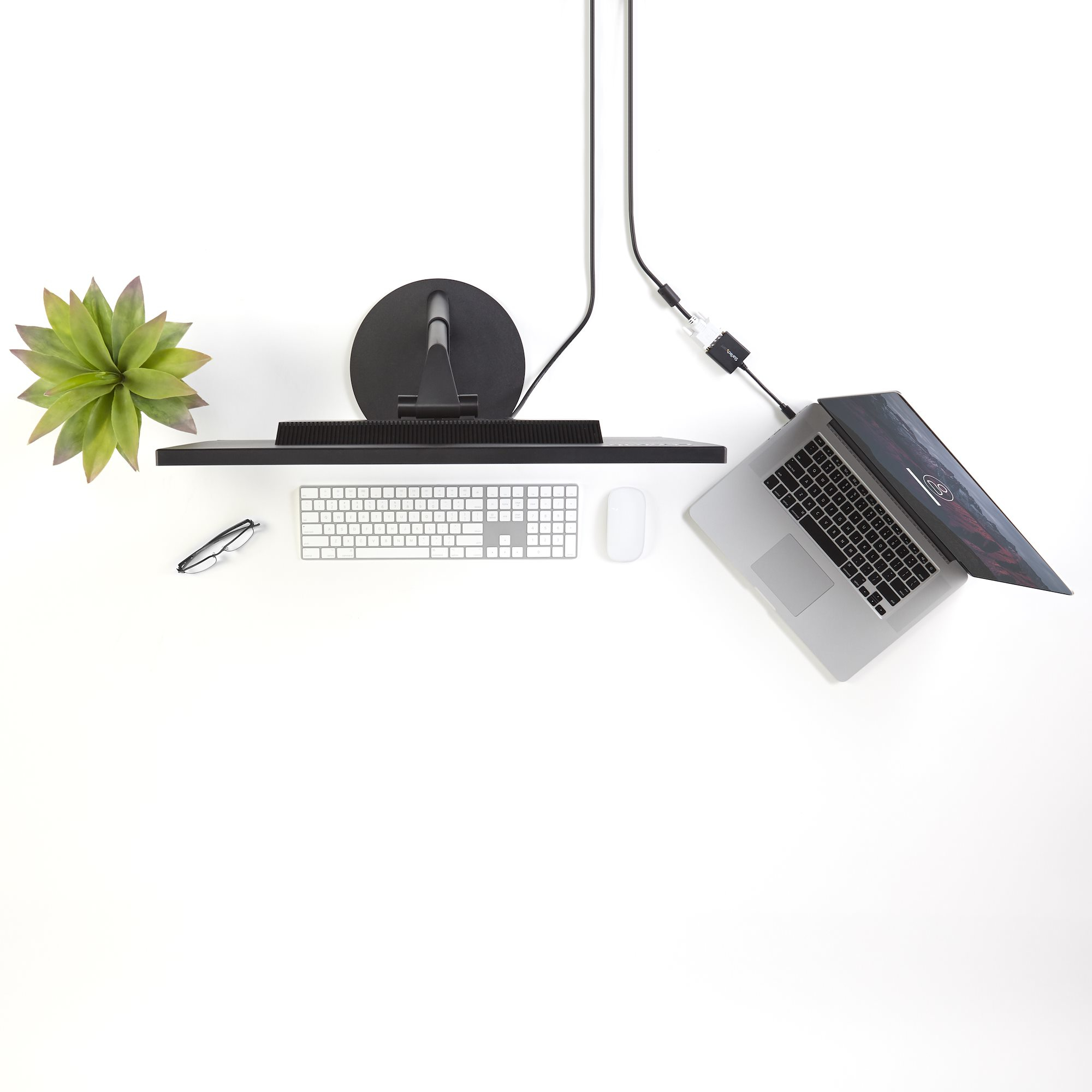 Vista cenital de un escritorio blanco con monitor, teclado, portátil, ratón, planta y gafas.