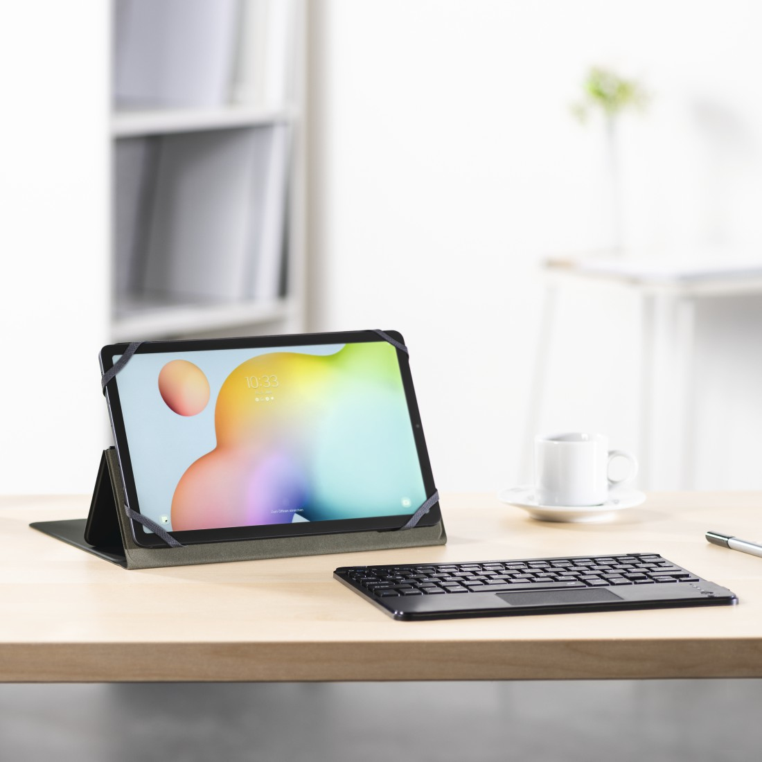Tablet mit Tastatur auf einem Holztisch. Eine Kaffeetasse und ein Stift sind ebenfalls vorhanden. Der Hintergrund zeigt einen weißen Schrank.