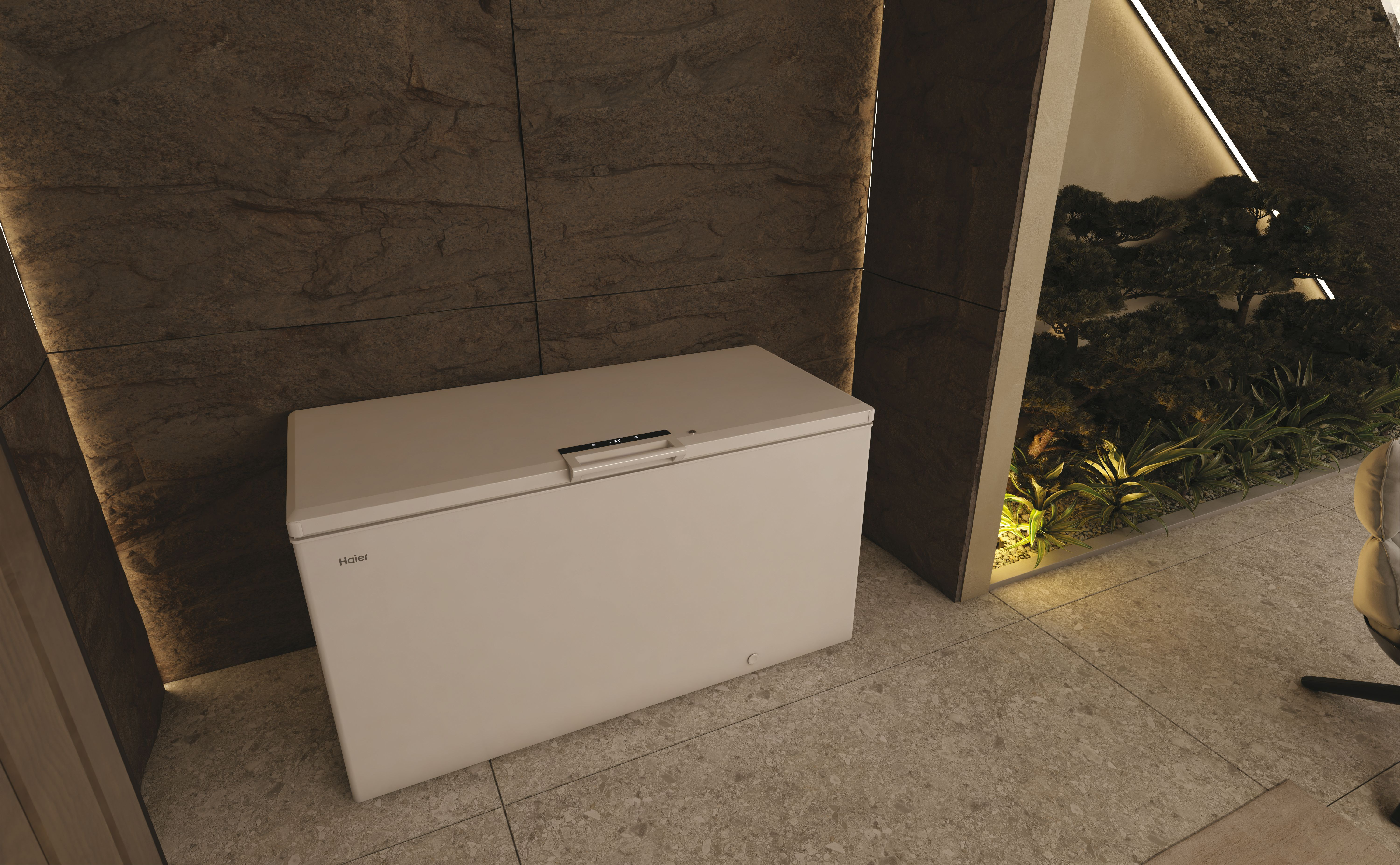 Un congelatore bianco Haier è vicino a un muro di pietra e a un giardino. Il coperchio del congelatore ha una maniglia.