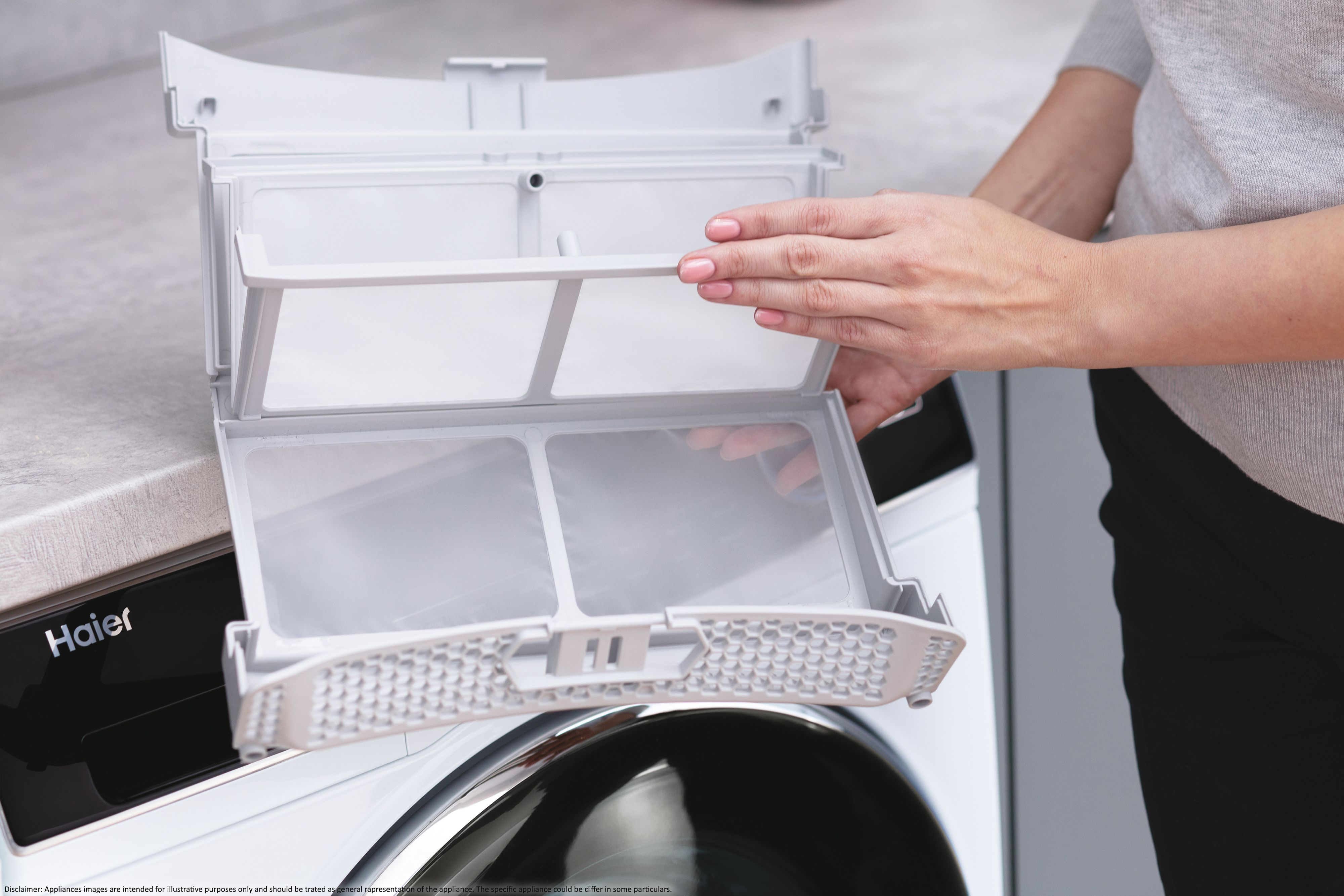 Osoba trzyma biały filtr z pralki Haier. Widoczne są różowe paznokcie.