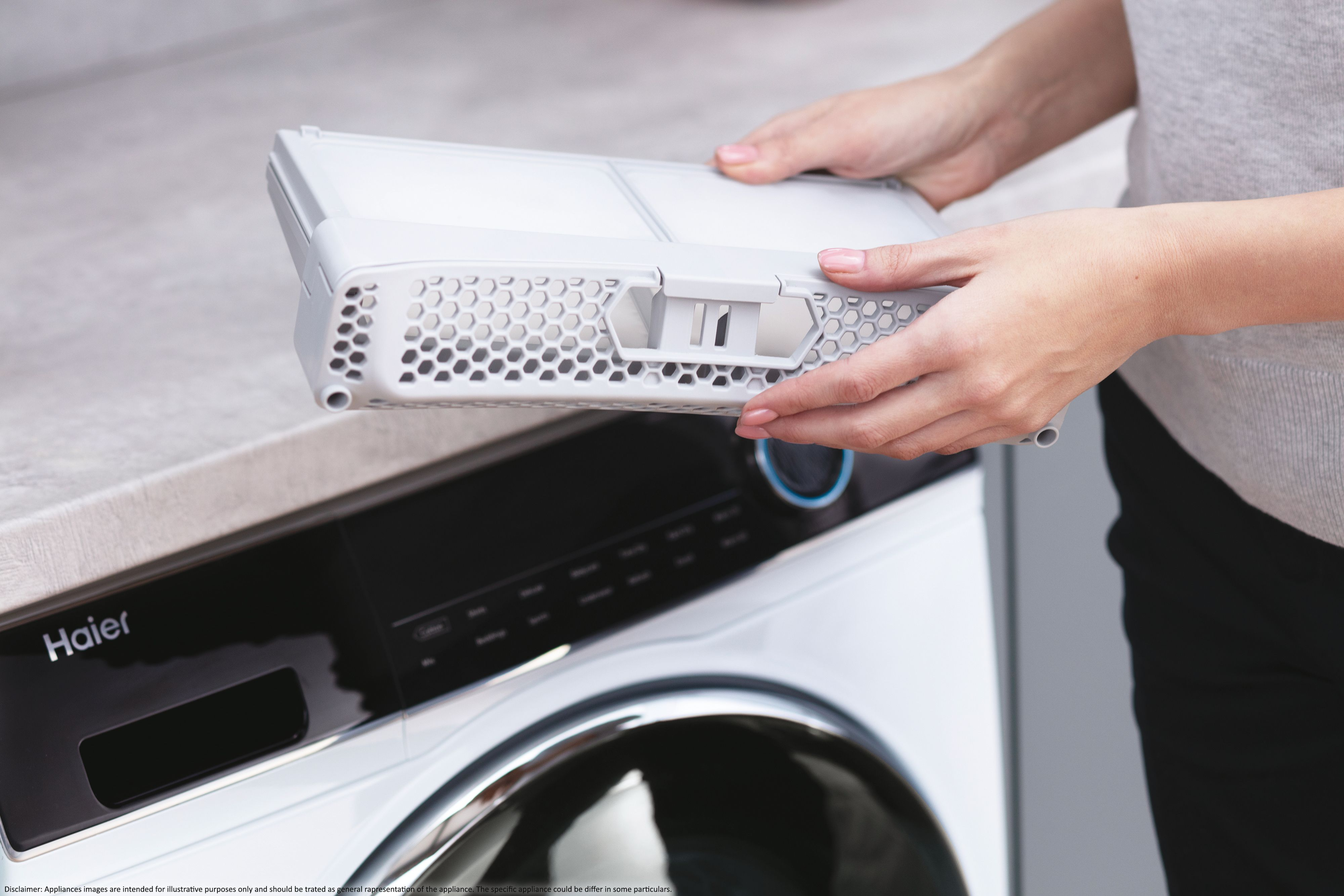 Una persona tiene in mano un filtro grigio di una lavatrice Haier. La macchina è bianca con un pannello nero.
