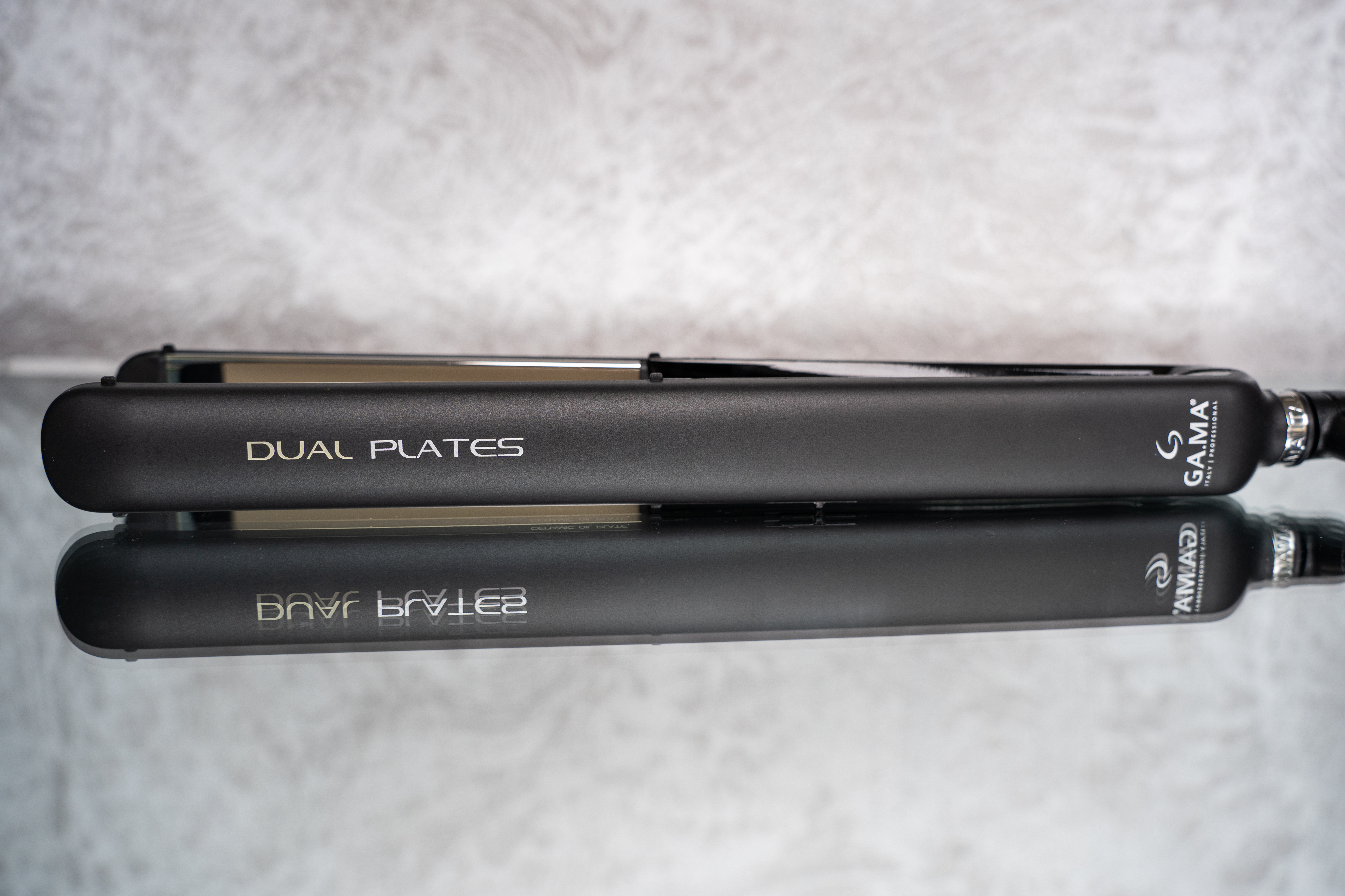 Piastra per capelli nera con testo 'DUAL PLATES'. Riflessa su superficie a specchio. Sfondo grigio.