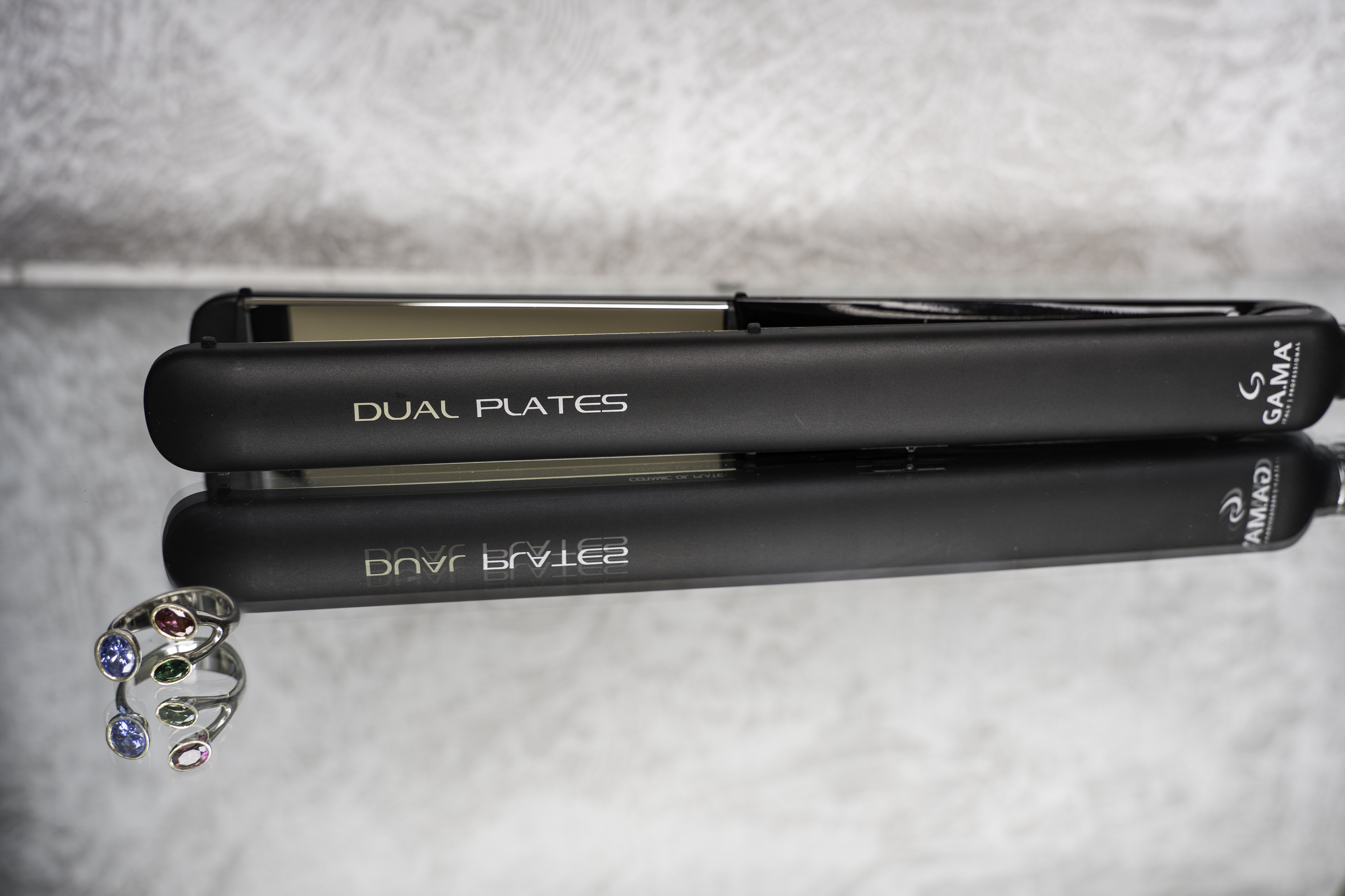 Piastra per capelli nera con testo 'DUAL PLATES'. Un anello con gemme colorate in primo piano. Riflesso speculare.