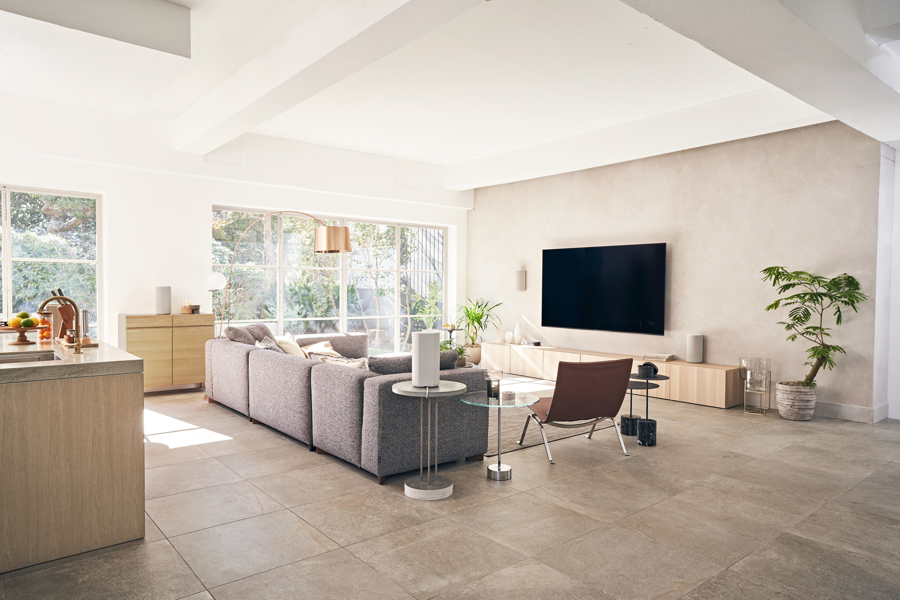 Wohnzimmer mit Sofa, TV und großen Fenstern. Enthält Pflanzen, Lampen und einen braunen Lederstuhl. Helles Farbschema.