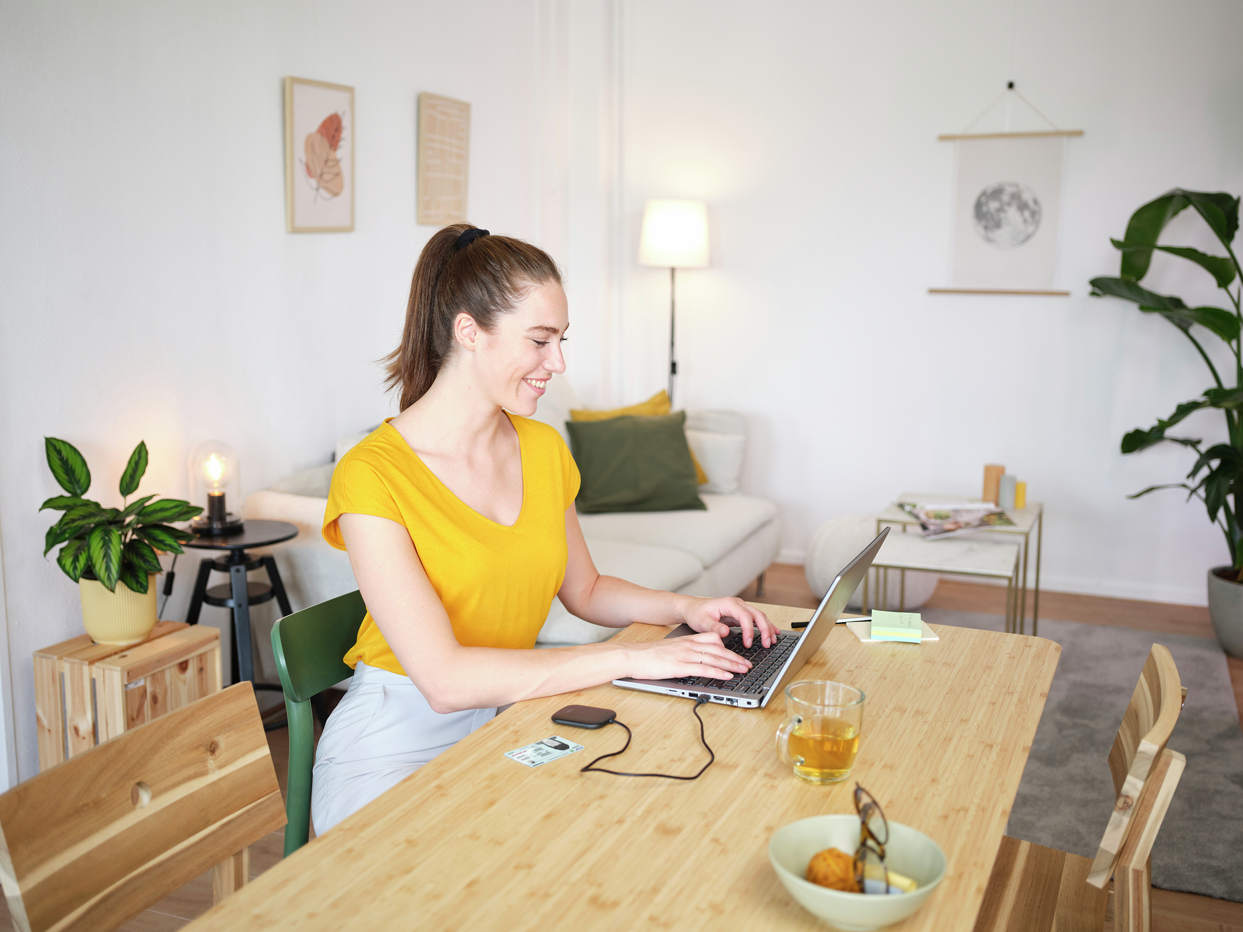 Una donna usa un laptop a un tavolo di legno. Ci sono tè, una ciotola e una pianta. Un divano e arte murale sullo sfondo.