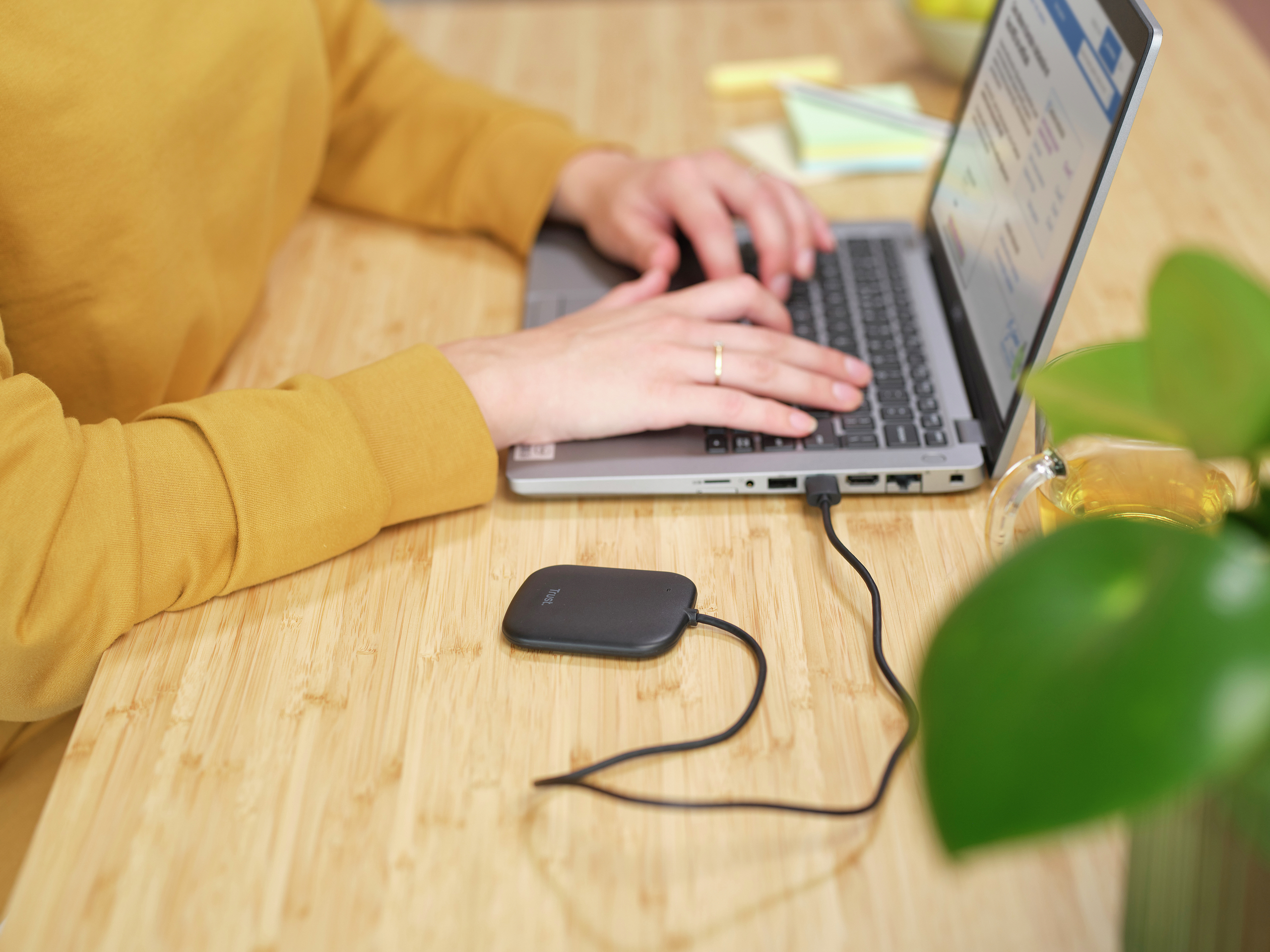 Persona che digita su un laptop, collegato a un dispositivo nero. Una tazza di tè e una pianta verde sullo sfondo.