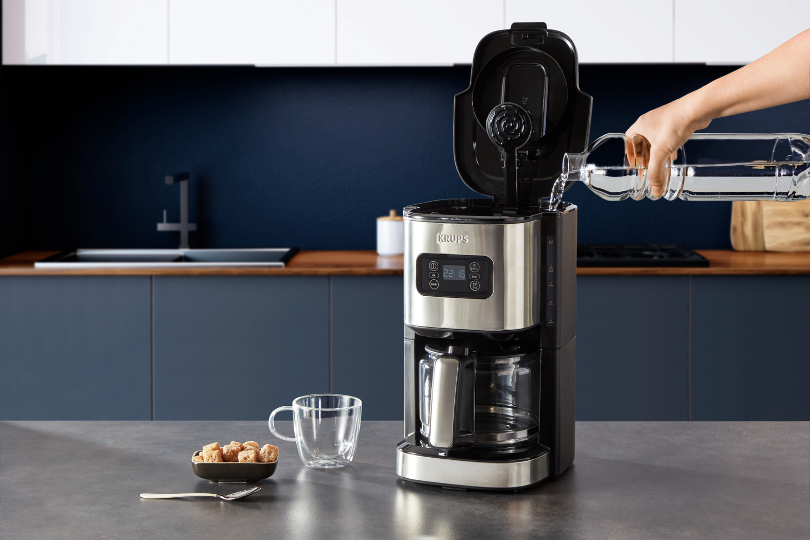 Ekspres do kawy napełniany wodą. Na blacie filiżanka, kostki cukru i łyżeczka. Kuchnia w tle.