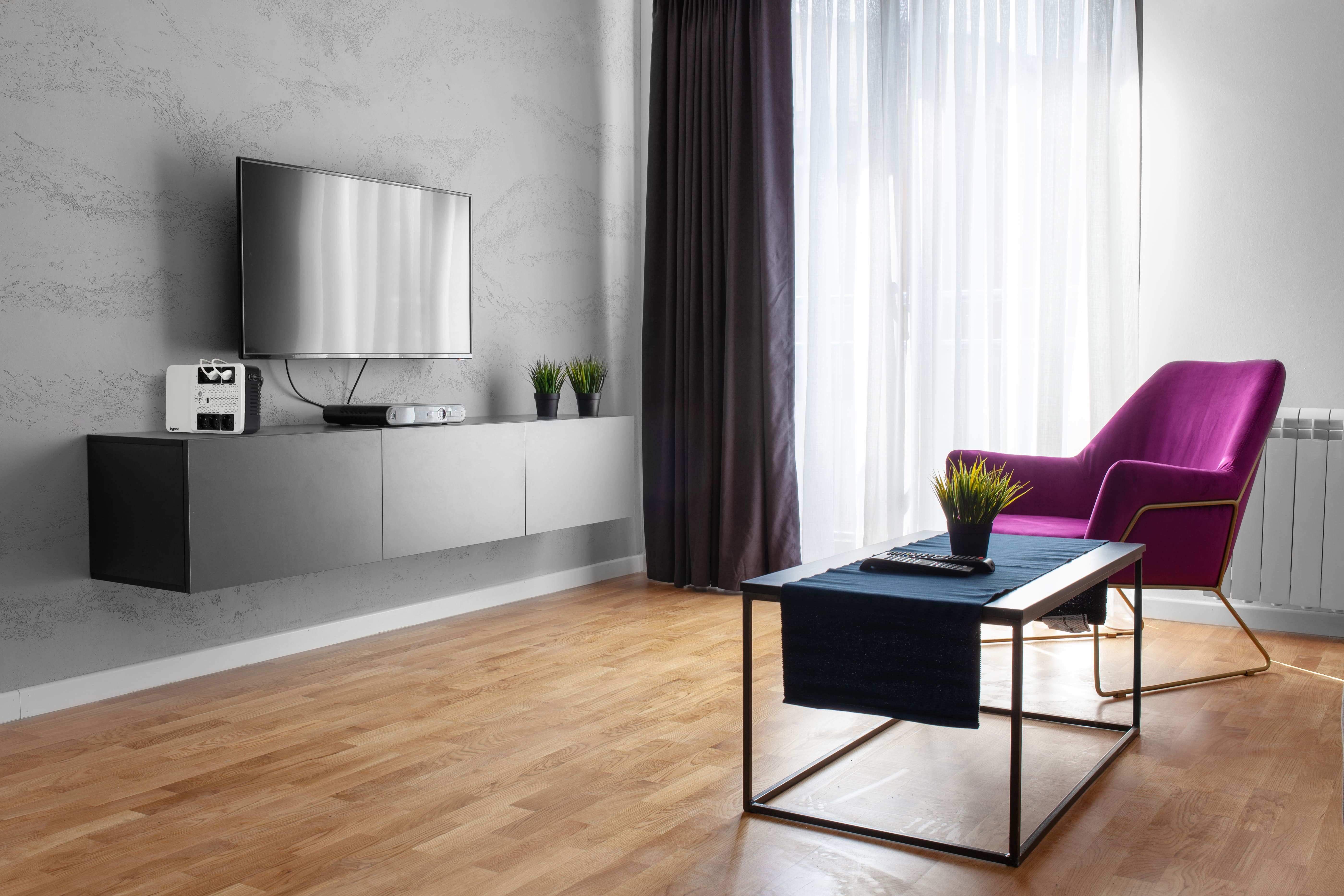 Modernes Wohnzimmer mit Fernseher, lila Stuhl, Couchtisch und Holzboden. Vorhänge und Sonnenlicht.
