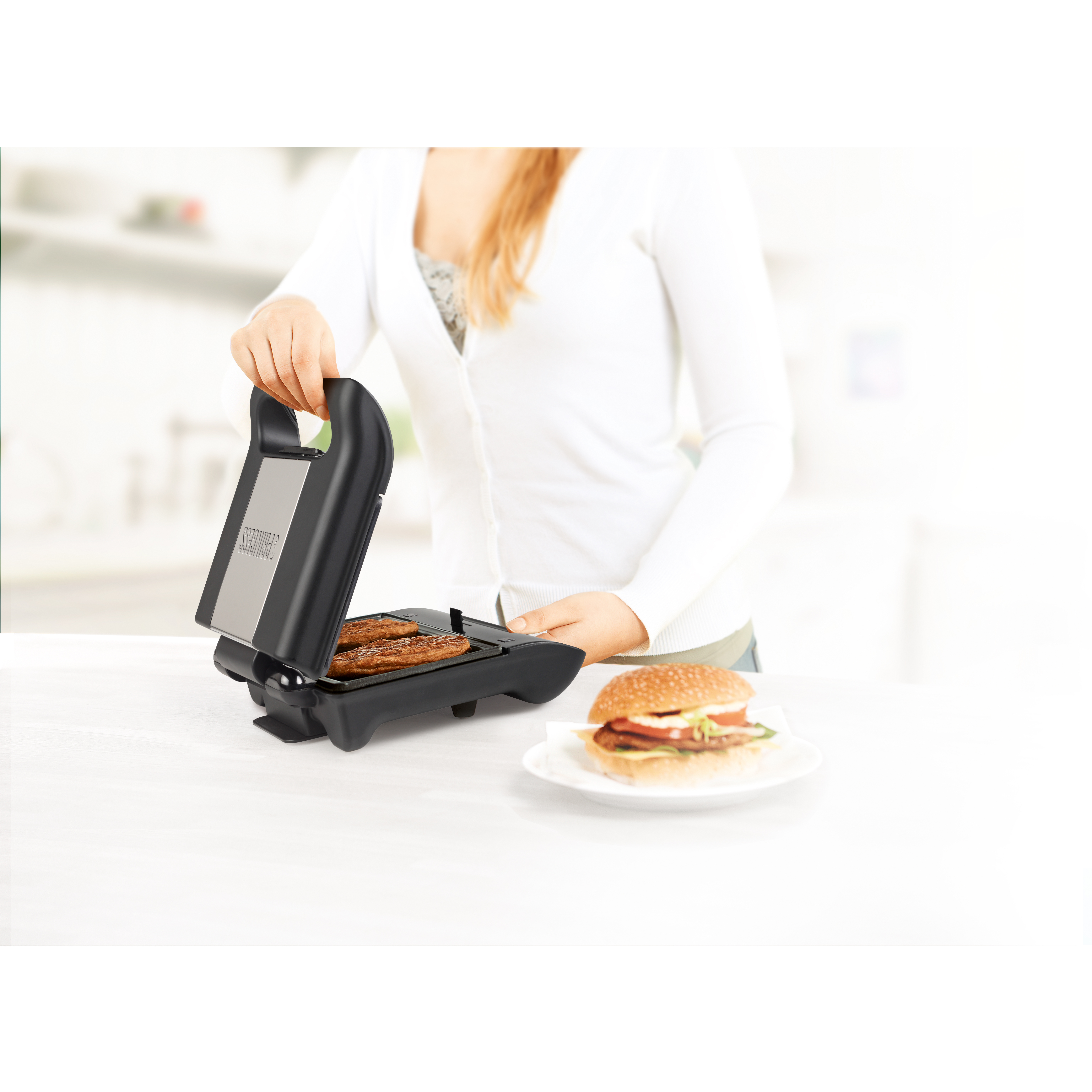 Una donna usa una macchina per sandwich. Ci sono hamburger sulla macchina e su un piatto. Lo sfondo è sfocato.
