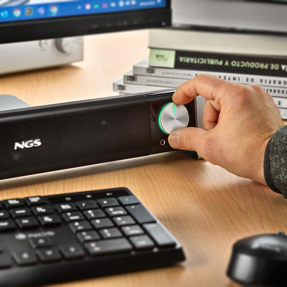 Una mano regola la manopola del volume su una soundbar nera. Tastiera, mouse e libri sono sulla scrivania di legno.