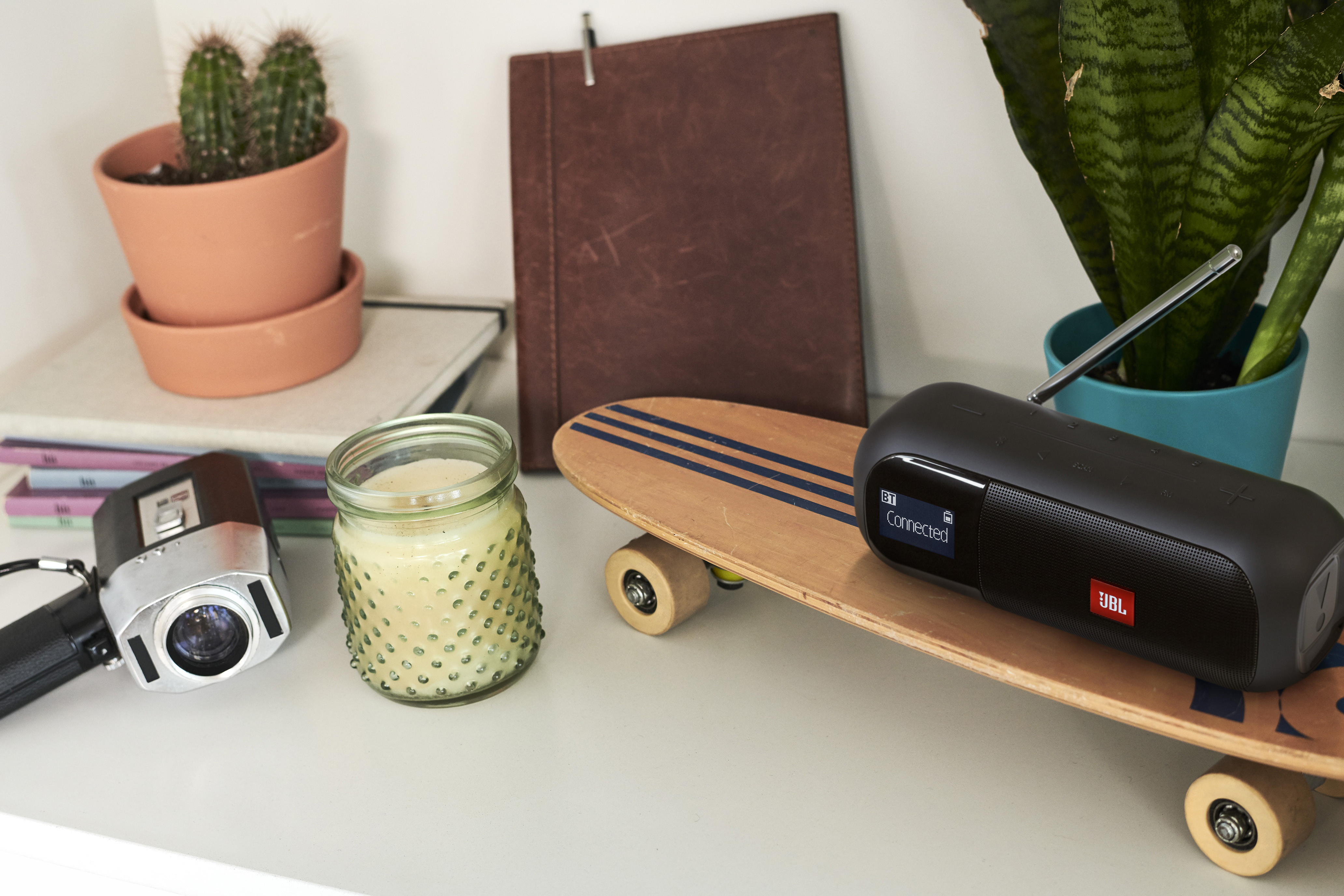 Natura morta con cactus, candela, skateboard, altoparlante JBL, videocamera e libri, su superficie bianca.