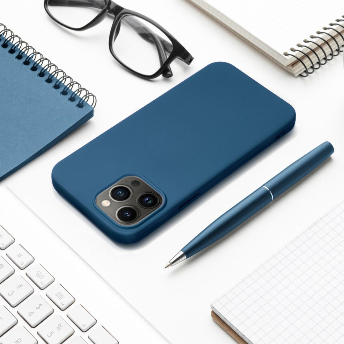 Eine blaue Handyhülle, ein Stift, ein Notizbuch, eine Brille und eine Tastatur auf einer weißen Oberfläche. Alles ist ordentlich angeordnet.