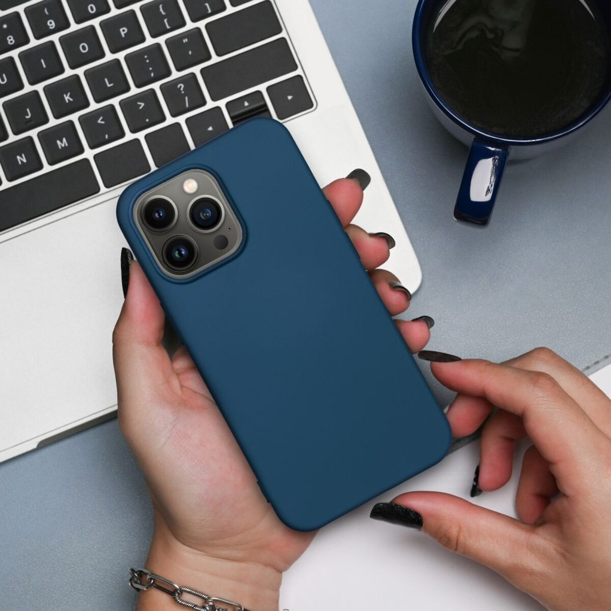 Eine Person hält ein blaues Telefon neben einem Laptop und einer Kaffeetasse. Die Nägel sind schwarz lackiert. Ein silbernes Armband ist zu sehen.