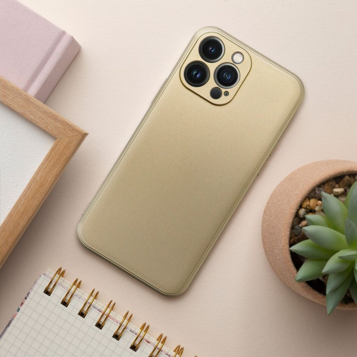 Ein goldenes Telefon mit mehreren Kameras liegt auf einer beigen Oberfläche, in der Nähe einer Pflanze, eines Notizblocks und eines Rahmens.
