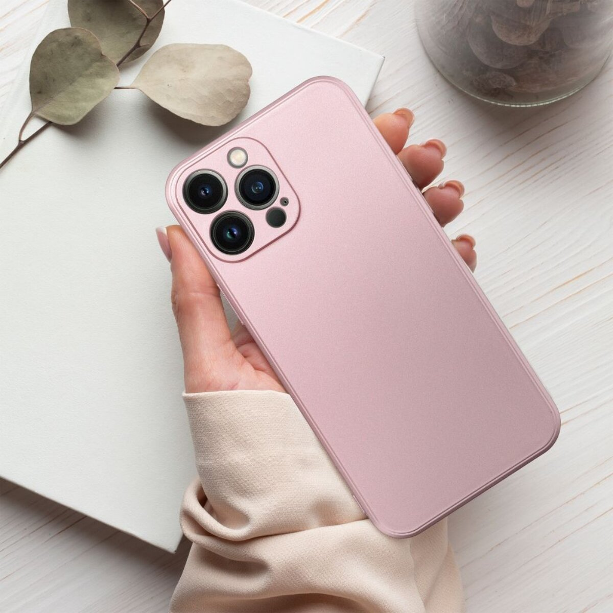 Eine Hand hält ein pinkfarbenes Telefon. Das Telefon hat drei Kameras. Der Hintergrund umfasst einen weißen Notizblock und ein Glas.