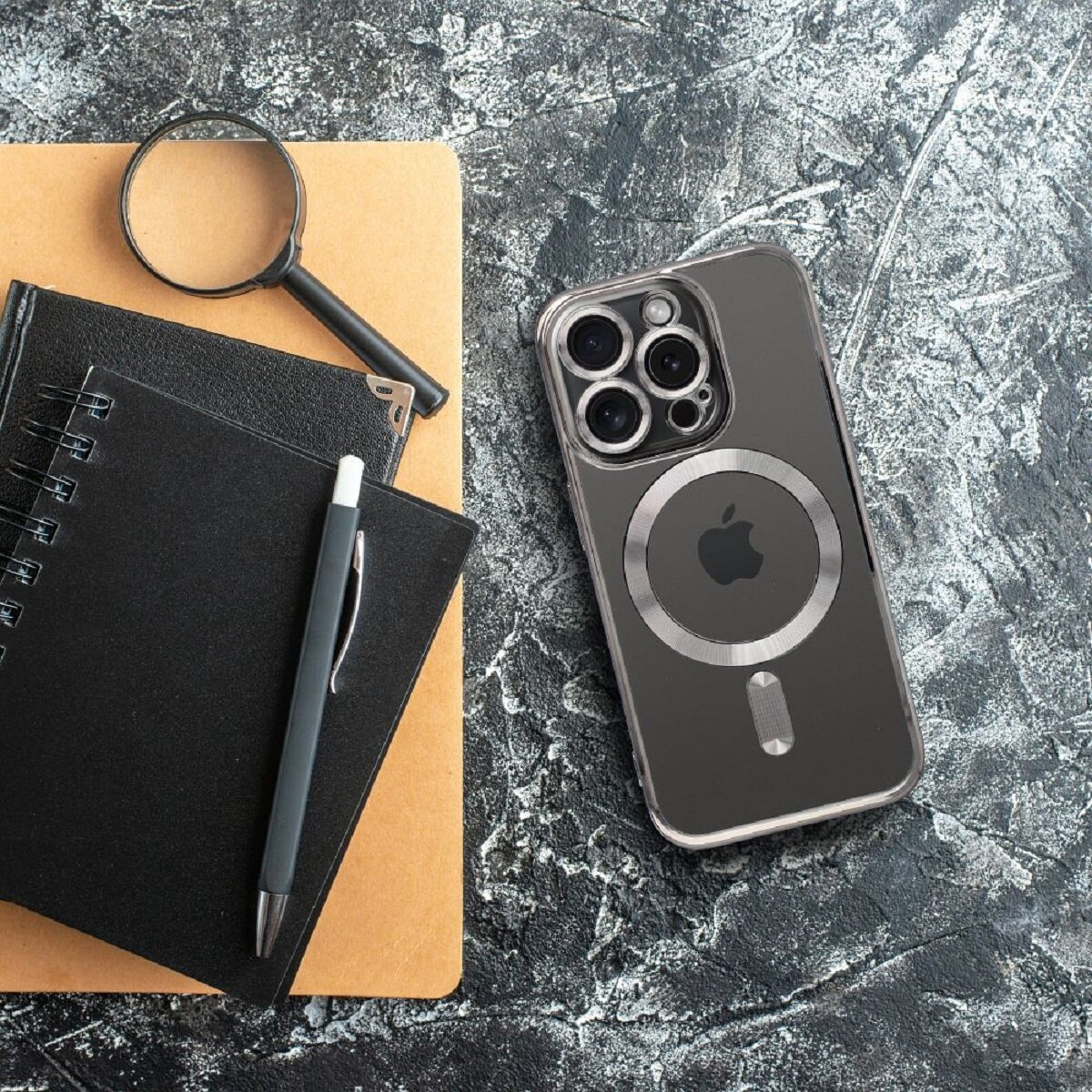 Ein Telefon in einer Hülle, Notizbücher, Stift und Lupe auf einer strukturierten Oberfläche. Das Telefon befindet sich in der Nähe der rechten Seite.