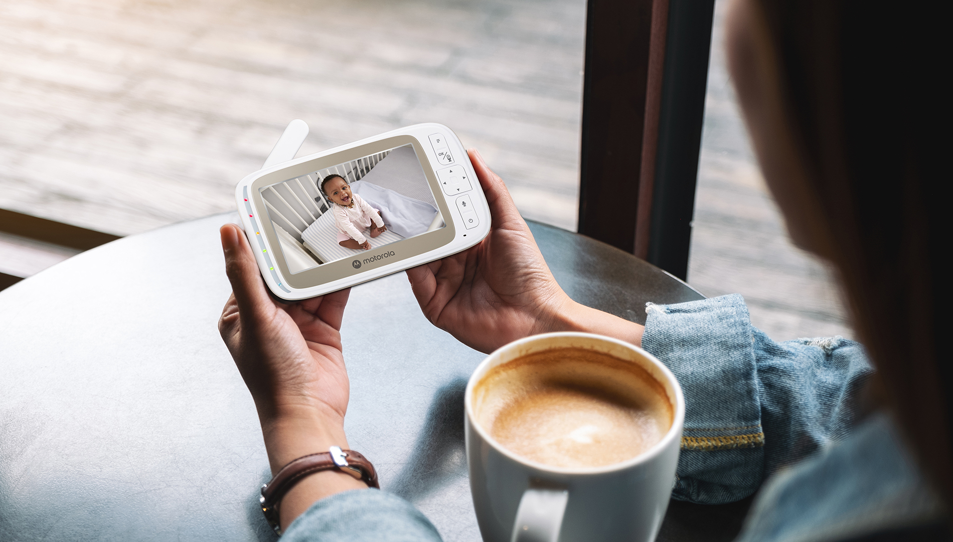 Eine Person hält einen Babyphone, das ein lächelndes Baby in einem Kinderbett zeigt. Eine Kaffeetasse steht im Vordergrund.
