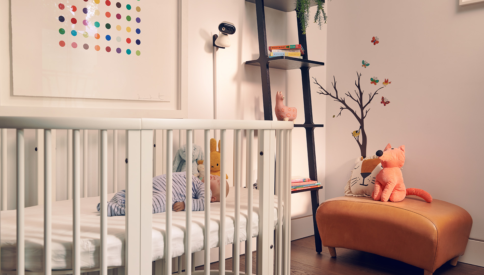 Een baby slaapt in een witte wieg. Een vossenknuffel zit op een leren poef. Muurkunst en een ladderplank staan in de buurt.
