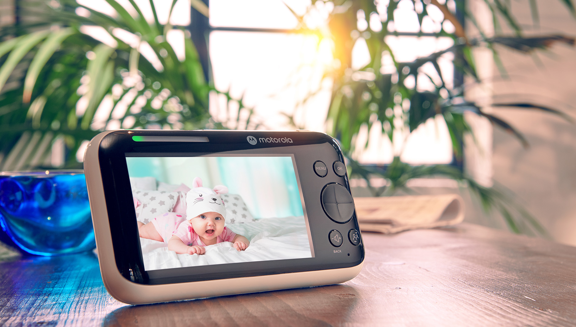 Een Motorola babyfoon toont een baby met een konijnenmuts. De monitor staat op een houten tafel. Een blauw glas en een plant staan op de achtergrond.