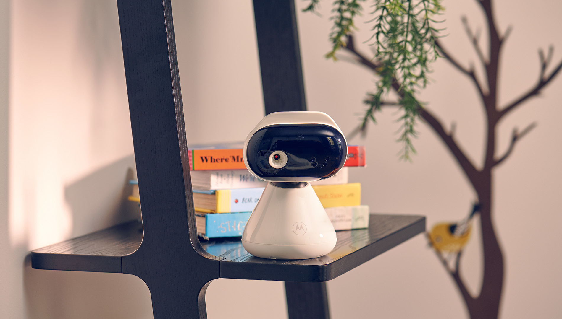 Ein weiß-schwarzer Babyphone steht auf einem Regal mit Büchern. Ein Baum schmückt die Wand dahinter.
