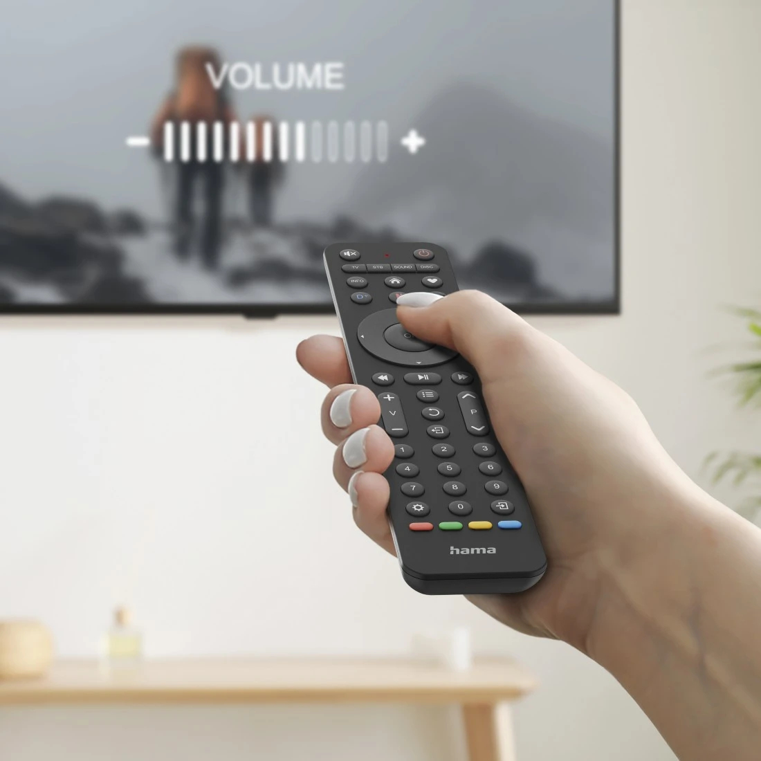 Eine Hand hält eine schwarze Fernbedienung und stellt die Lautstärke am Fernseher ein. Der Bildschirm zeigt ein verschwommenes Bild.