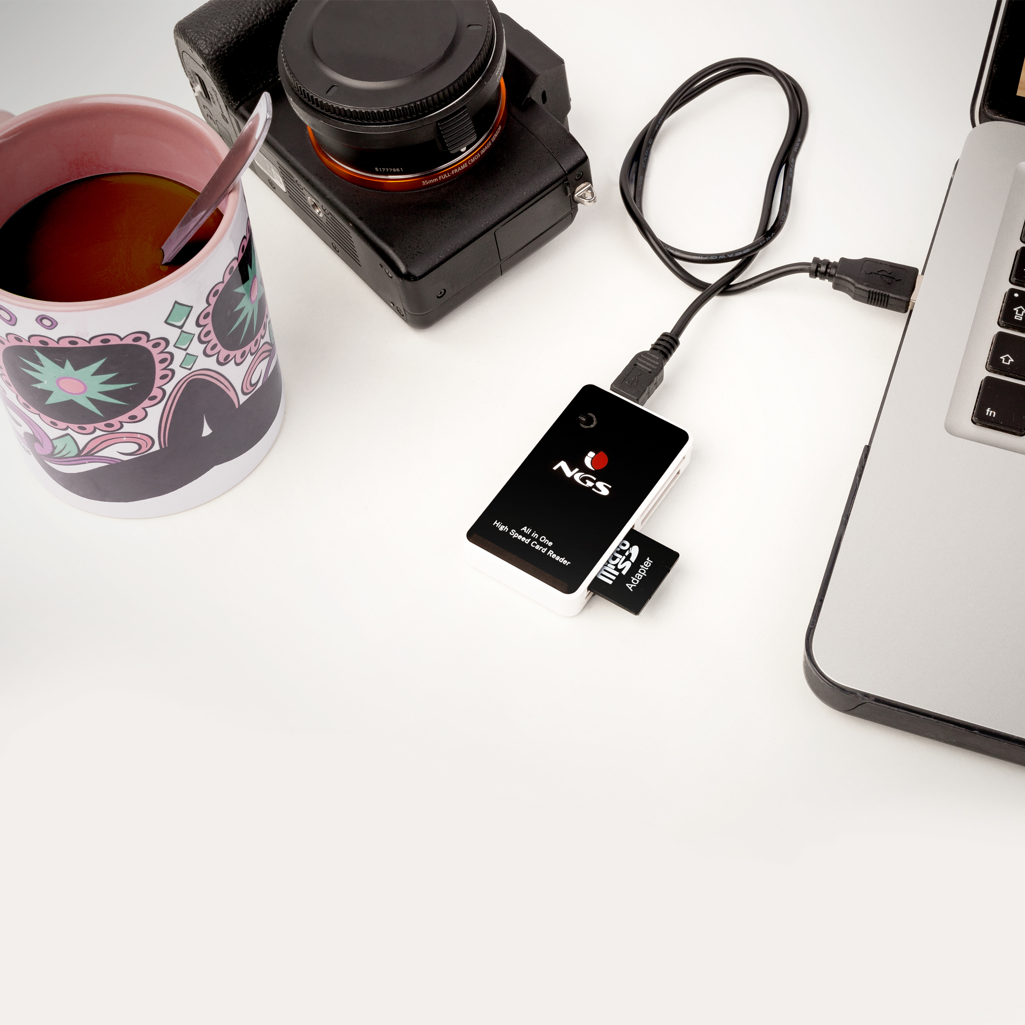 Escena de escritorio con cámara, taza, portátil y lector de tarjetas. Cable USB conecta el lector al portátil.
