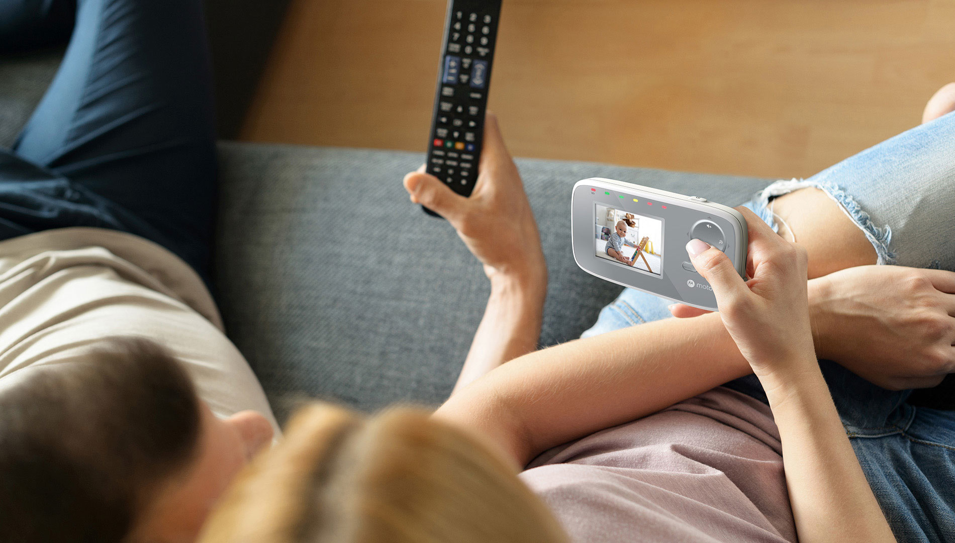 A person holds a baby monitor, watching a baby. Another person holds a remote. Both are seated on a gray couch.