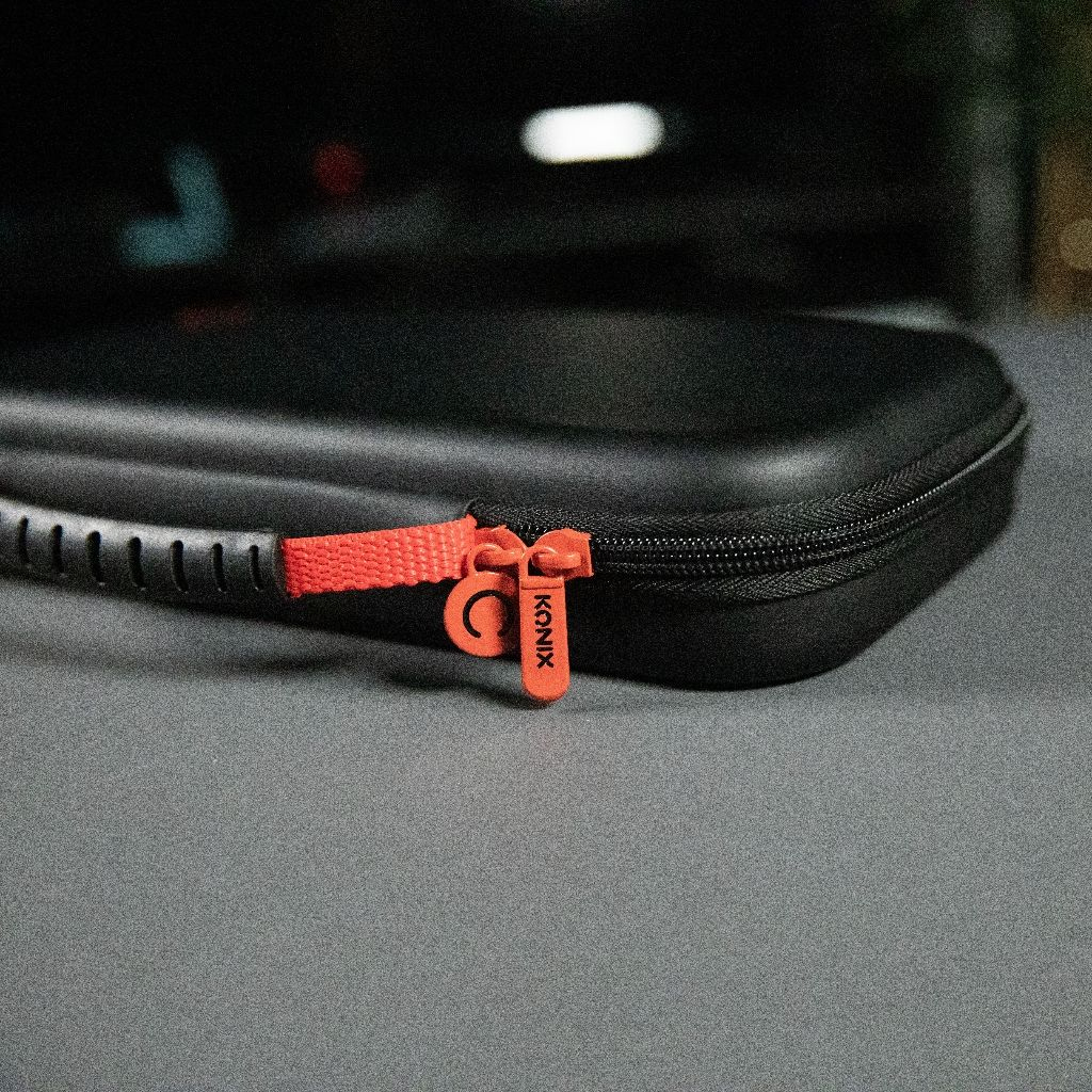 Black case with orange zipper. The zipper pull reads 'KONIX'. The case is on a gray surface with blurred background details.