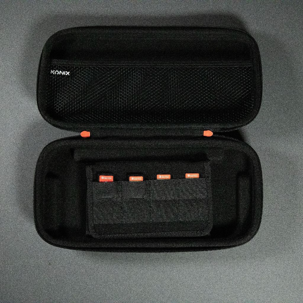 Open black KONIX case with orange accents. Inside, a mesh pocket and a holder with game cartridges are visible.