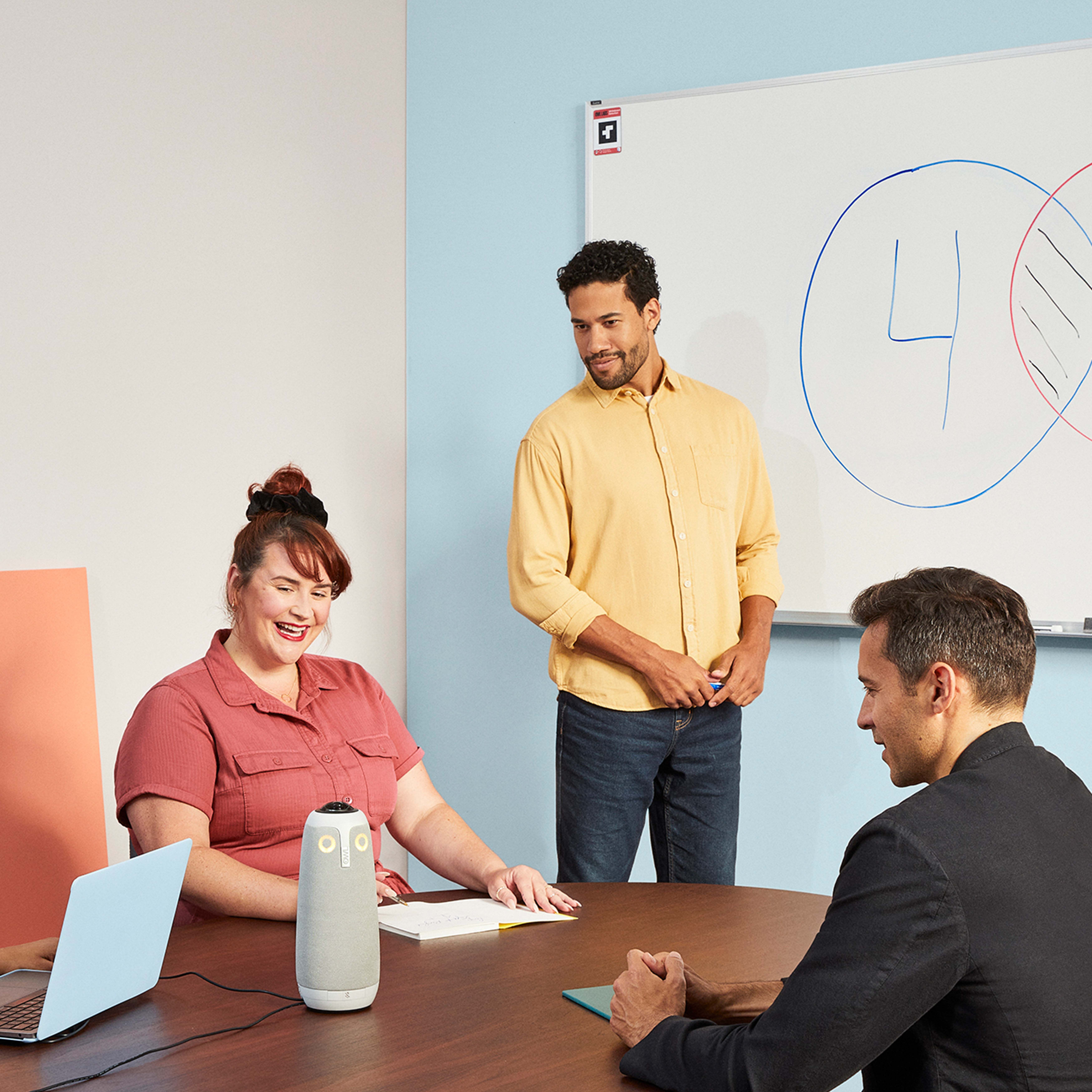 Drei Personen in einem Meeting. Eine Tafel mit Diagrammen, ein Lautsprecher und ein Laptop auf einem braunen Tisch.