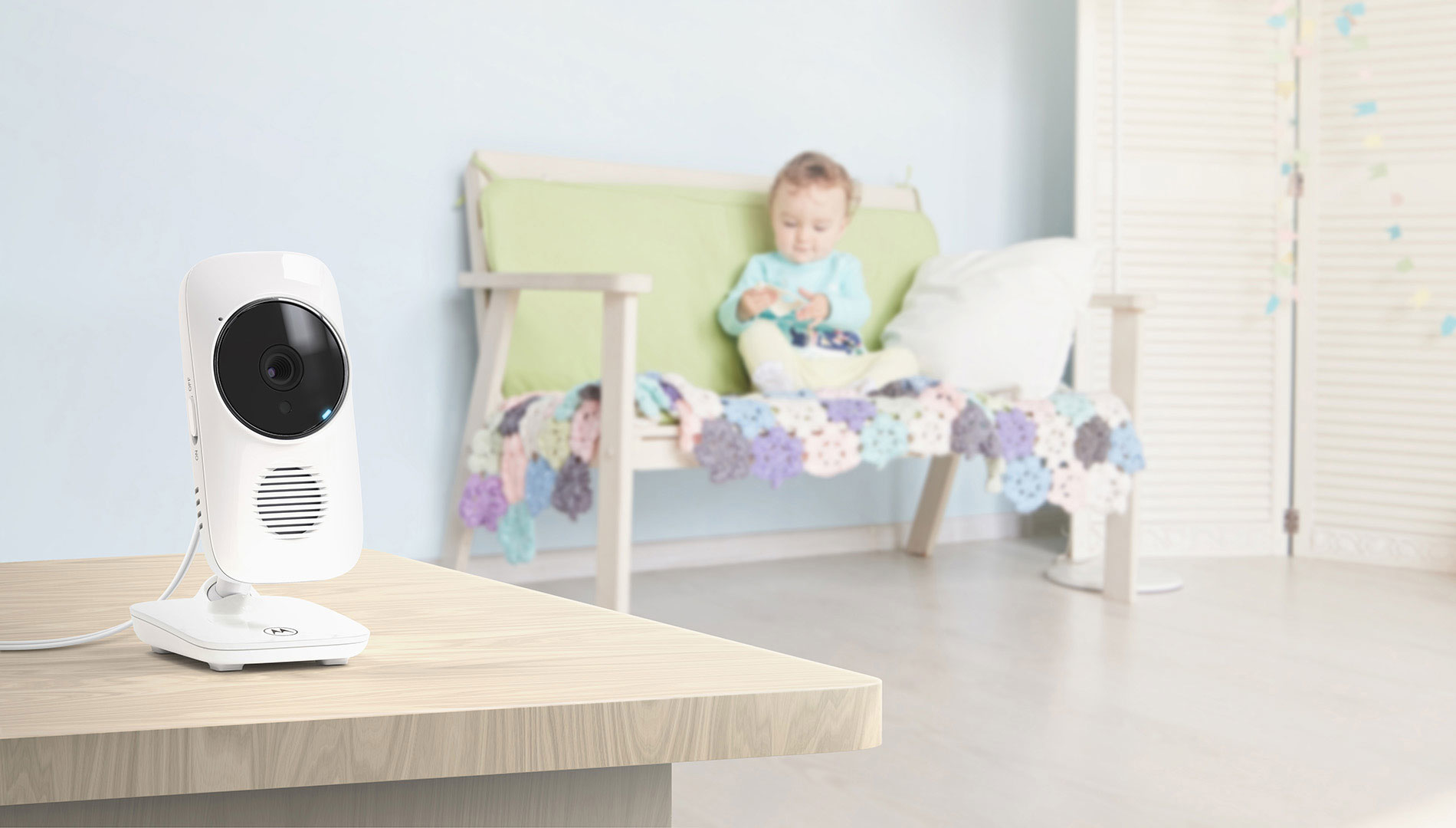Ein Babyphone auf einem Tisch, im Hintergrund sitzt ein Baby auf einer Couch.