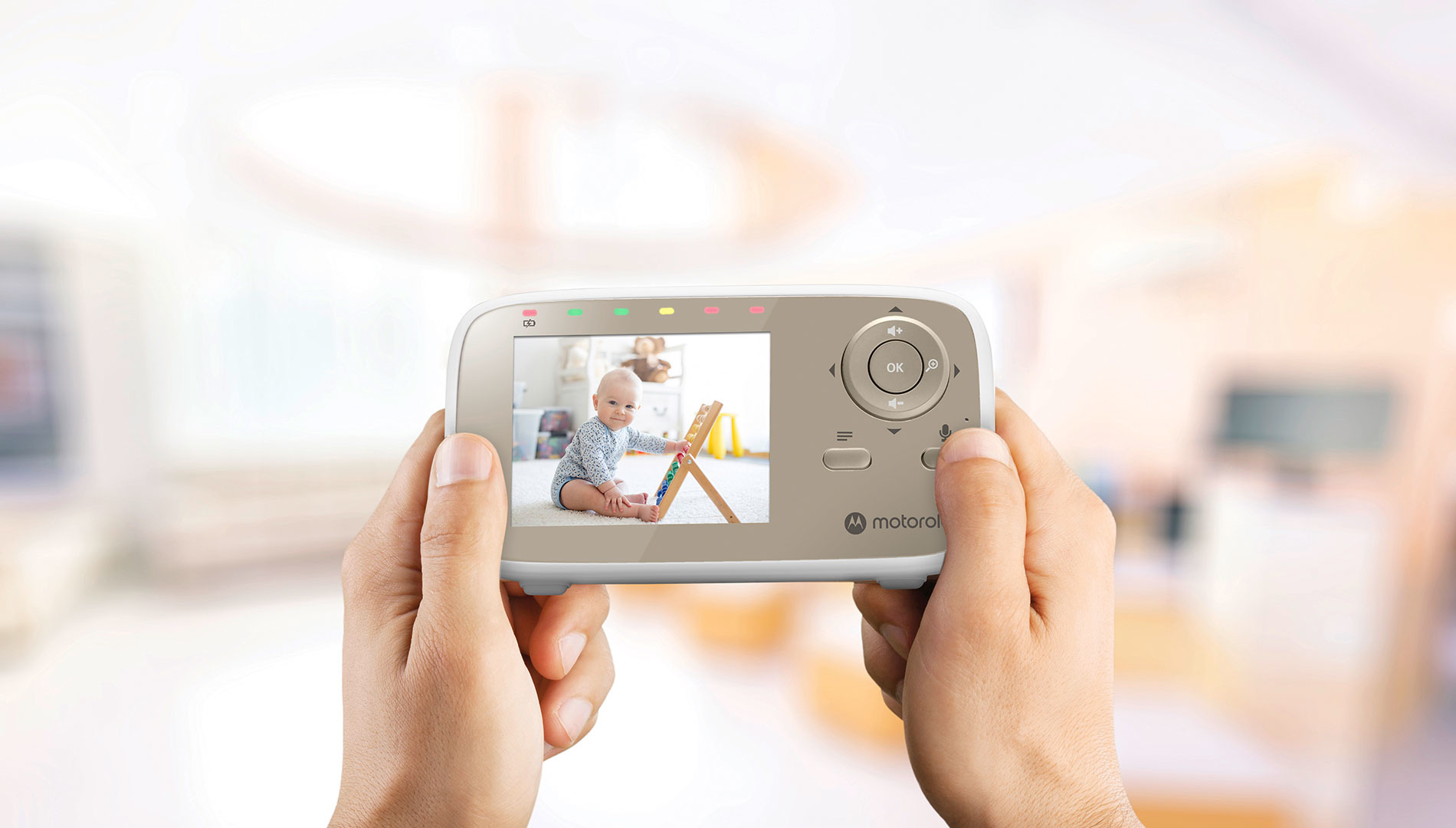 Hände halten einen Babyphone-Monitor, der ein Baby beim Spielen mit einem Spielzeug zeigt. Der Hintergrund ist ein verschwommener Raum.