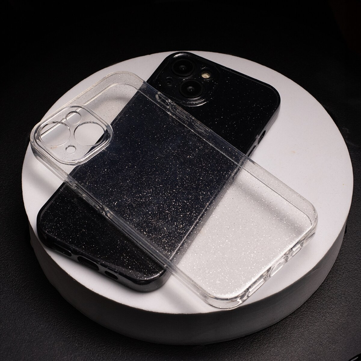 Ein schwarzes Smartphone mit einer glitzernden Hülle und einer transparenten Hülle auf einer weißen, kreisförmigen Plattform vor schwarzem Hintergrund.