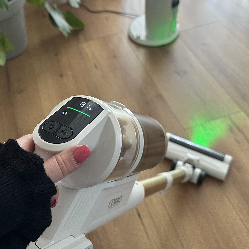 Ręka trzyma biało-złoty odkurzacz. Odkurzacz znajduje się na drewnianej podłodze, z zielonym światłem wydobywającym się z jego głowy.