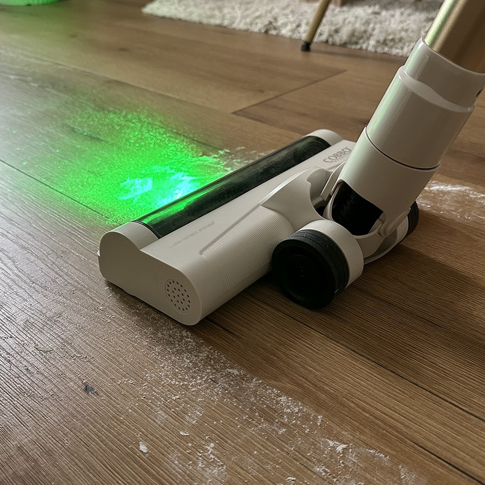 Biała głowica odkurzacza czyści drewnianą podłogę. Zielone światło lasera oświetla kurz. Dywan i noga krzesła w tle.