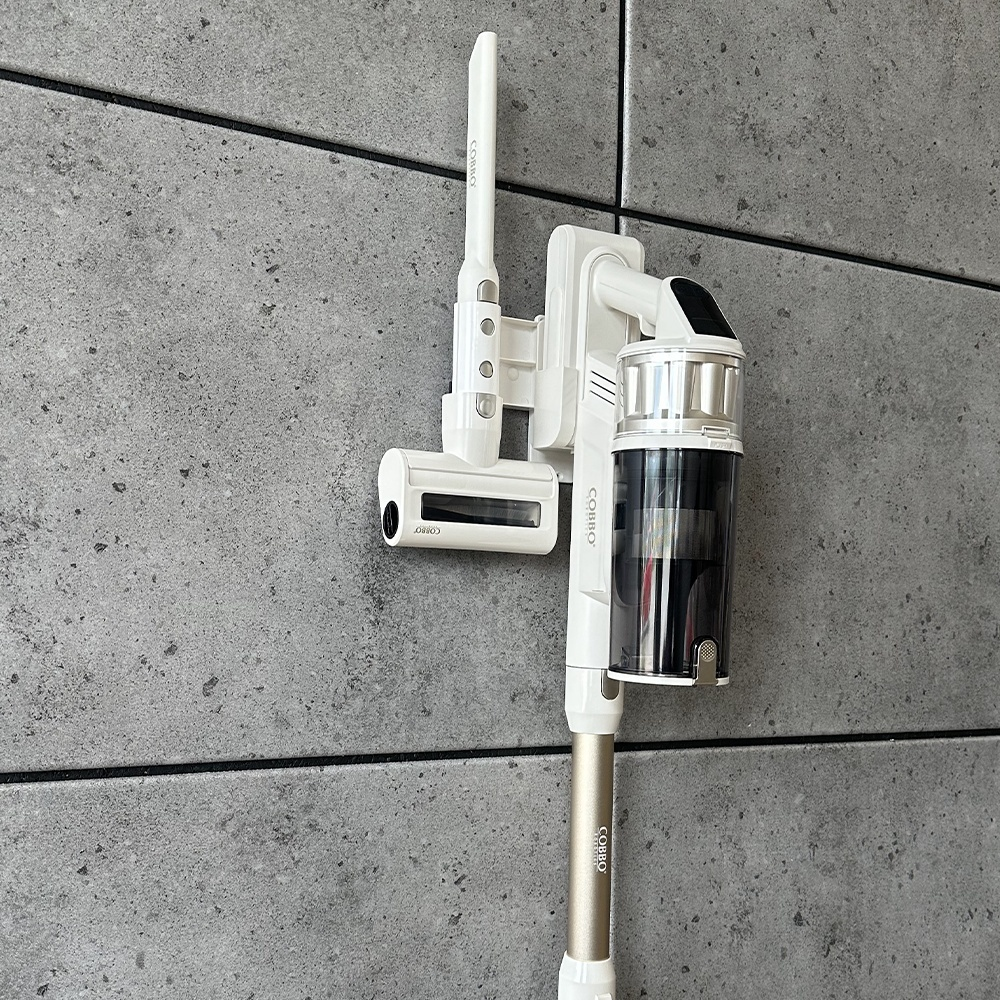 Biało-złoty odkurzacz COBBO jest zamontowany na szarej ścianie. Odkurzacz stoi pionowo, pokazując pojemnik na kurz i uchwyt.
