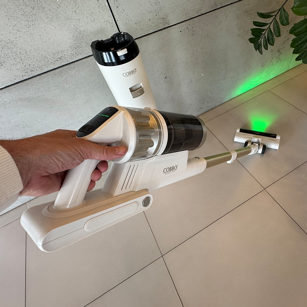 Biało-czarny odkurzacz bezprzewodowy trzymany przez osobę. Odkurzacz ma zielone światło. Tło to podłoga wyłożona kafelkami.