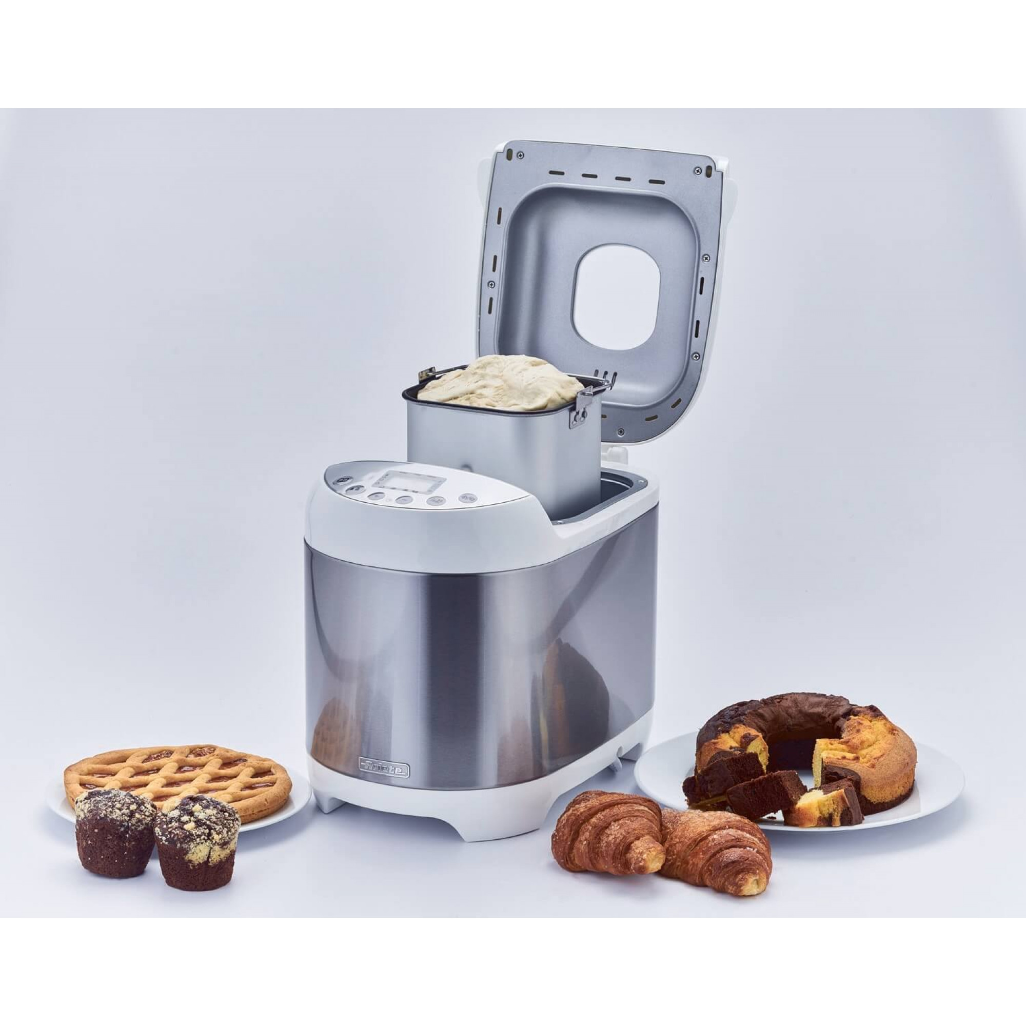 Brotbackautomat mit offenem Deckel, verschiedene Backwaren auf weißer Oberfläche: Kuchen, Croissant, Muffins und Kuchen.