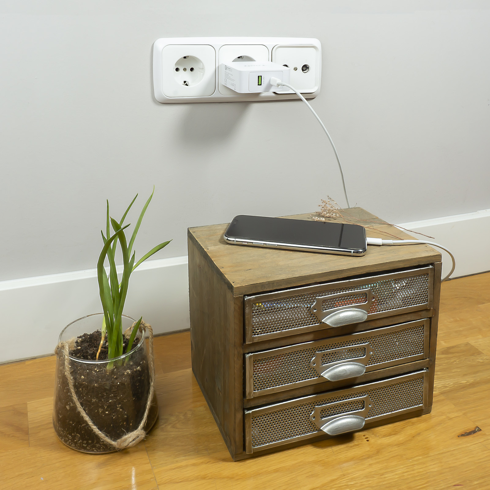Un teléfono cargando en una mesita de noche de madera. Una planta en una maceta de cristal está a la izquierda. Enchufe en la pared.