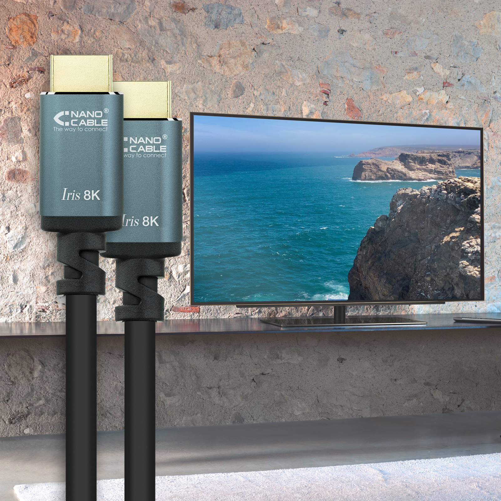 Dos cables HDMI con conectores dorados. Un televisor muestra un paisaje costero. Cables grises frente a una pared de piedra.