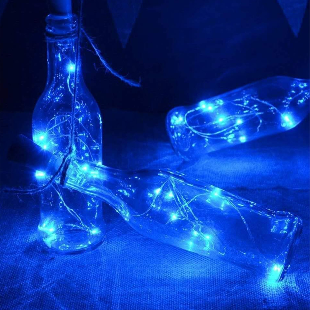 Drei Glasflaschen mit blauen Feenlichtern. Zwei Flaschen liegen, eine steht. Alle stehen auf einer blauen Oberfläche.