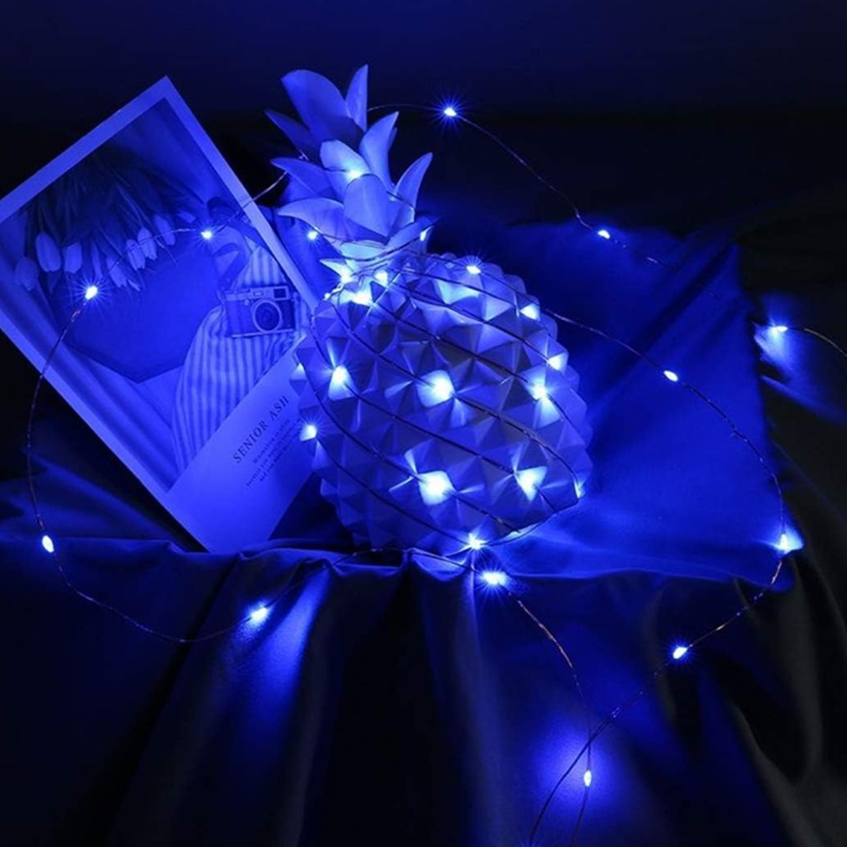 Eine ananasförmige Dekoration mit blauen Lichtern neben einem Buch. Die Szene ist in blaues Licht getaucht. Dunkler Hintergrund.