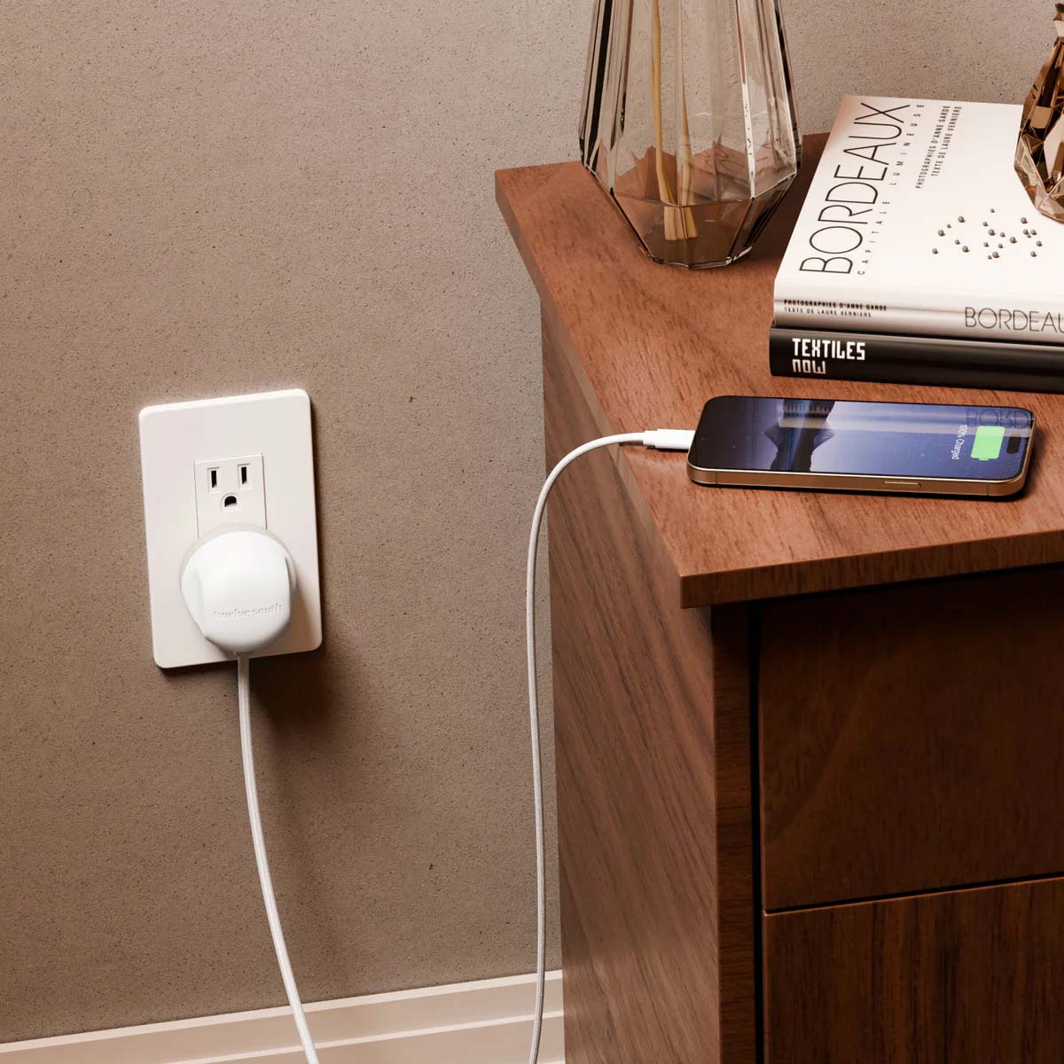 Ein Telefon, das auf einem Nachttisch geladen wird. Eine Steckdose mit Stecker. Bücher und ein dekoratives Glasobjekt sind sichtbar.