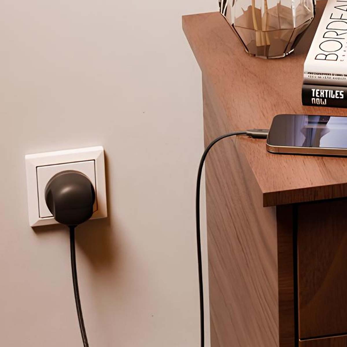 Ein schwarzer Stecker steckt in einer Steckdose. Ein Telefon wird auf einem hölzernen Nachttisch geladen. Bücher und eine Vase stehen auch auf dem Ständer.