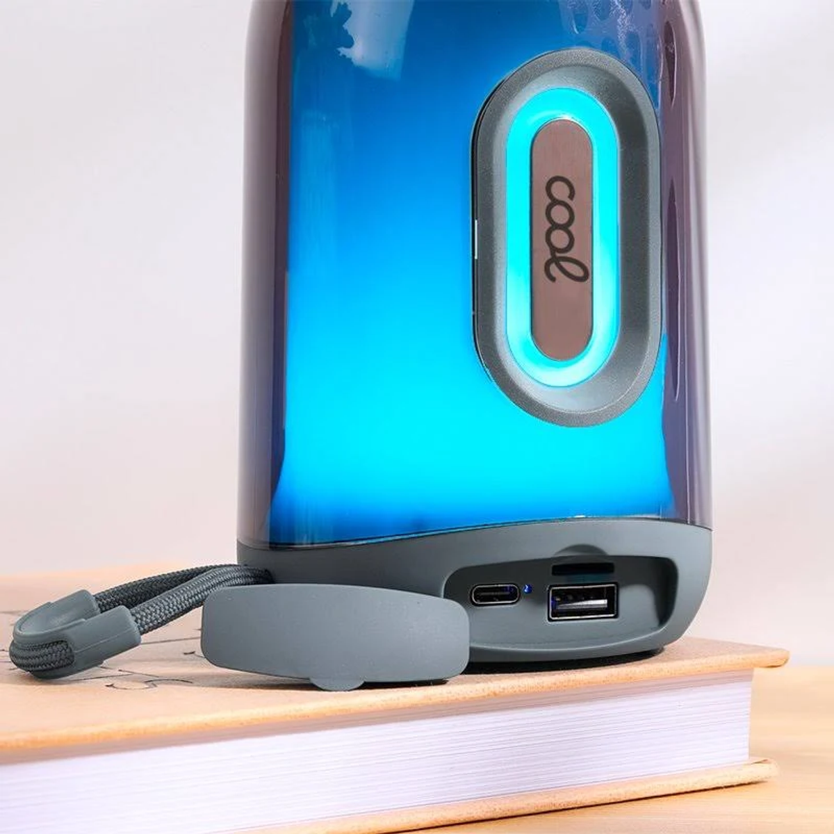 Un altavoz portátil azul iluminado con la etiqueta 'cool'. Se encuentra sobre un libro. Son visibles una correa gris y un puerto USB.