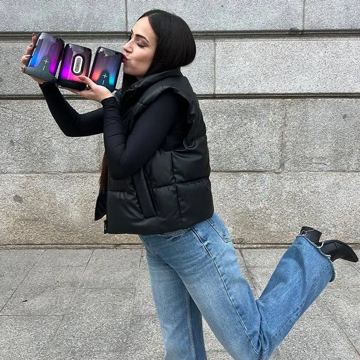 Una mujer besa tres altavoces con luces de colores. Lleva un chaleco negro, vaqueros y botas, de pie contra una pared de piedra.