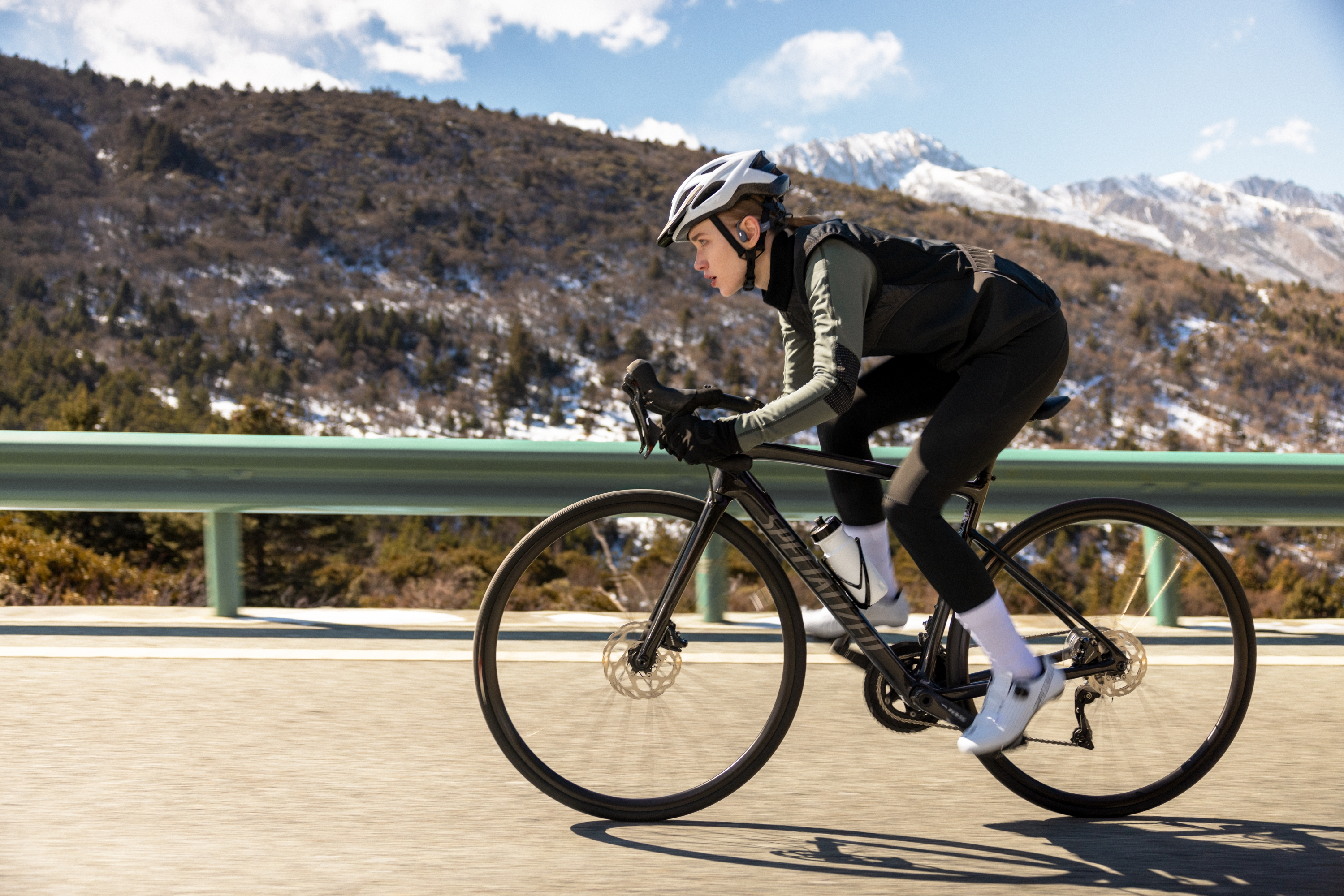 Un cycliste fait du vélo sur une route, montagnes en arrière-plan. Elle porte un casque et tenue sport.