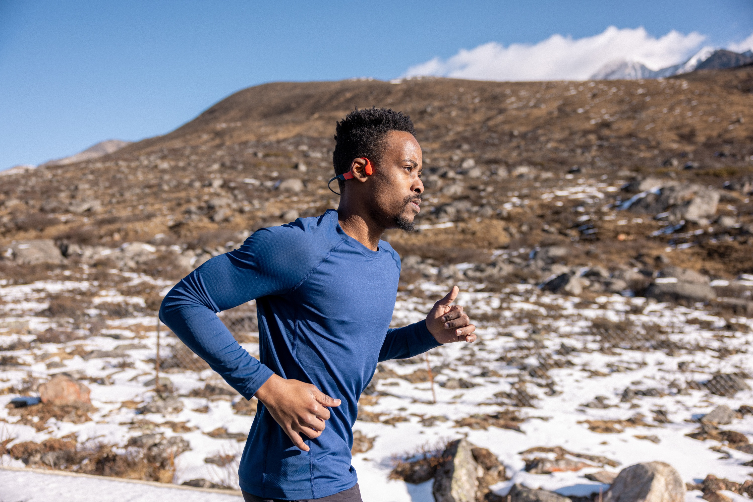 Un homme court en plein air en chemise bleue avec des écouteurs rouges. Montagnes et neige en arrière-plan.